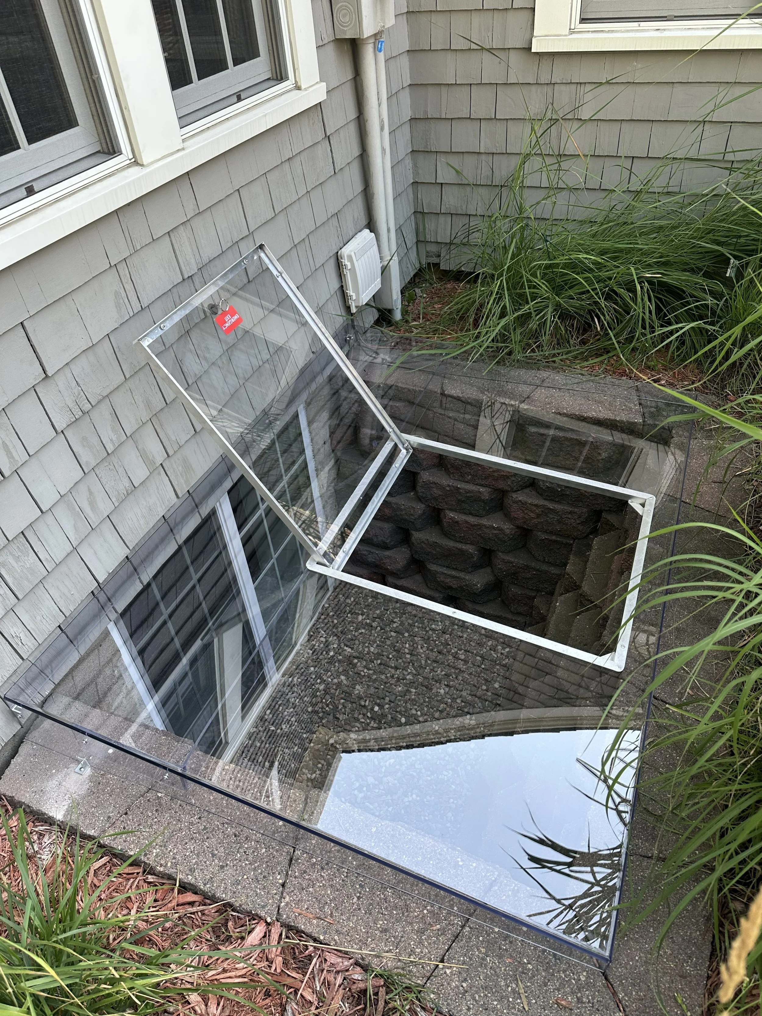 A glass box with a red 'danger' tag is placed outside near a house wall with gray siding, next to tall grass and plants, and a small drainage area with rocks, a window, and an electrical unit.