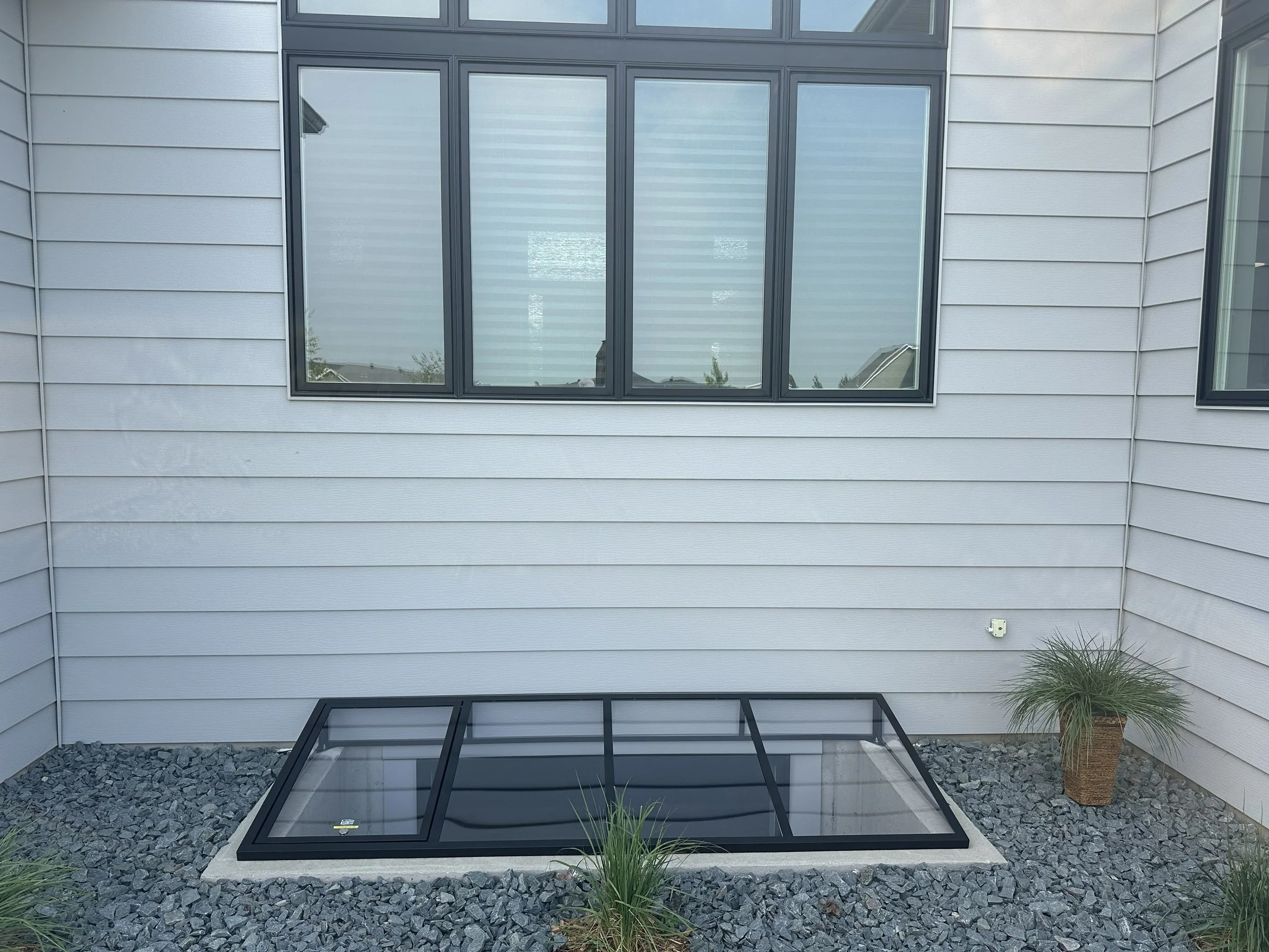 View of a modern house exterior with gray siding, large windows, and a small backyard enclosed by rock landscaping and a few plants.