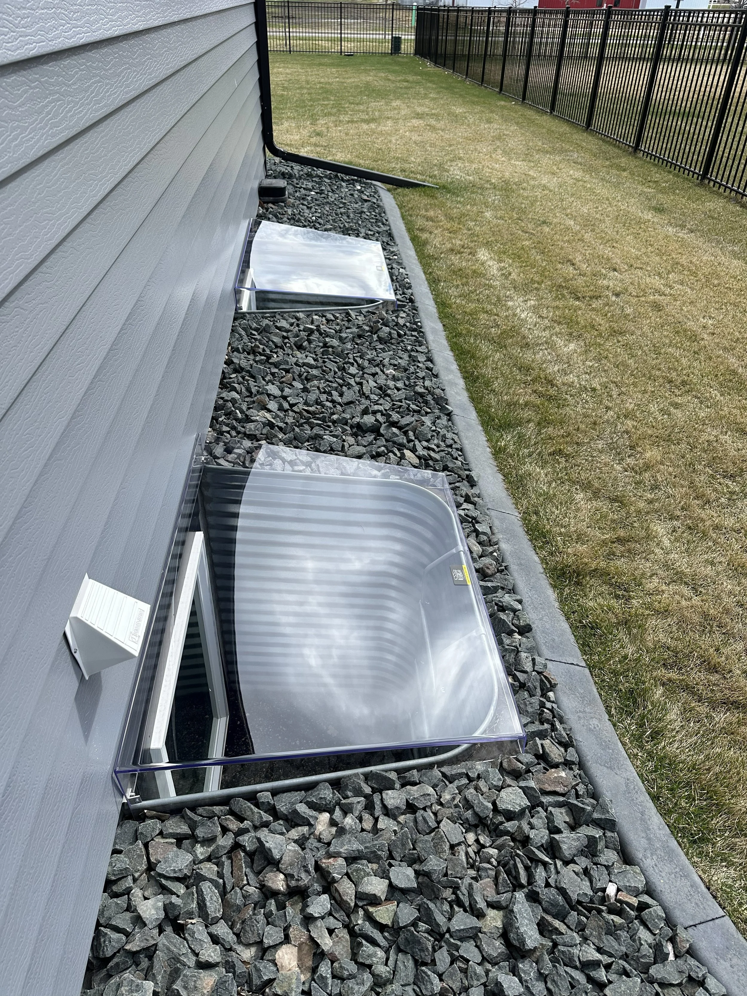 A backyard with a gray house, black metal fence, and green grass. There are three clear plastic pet carriers on a bed of gray rocks next to the house wall, with a window visible above the carriers.