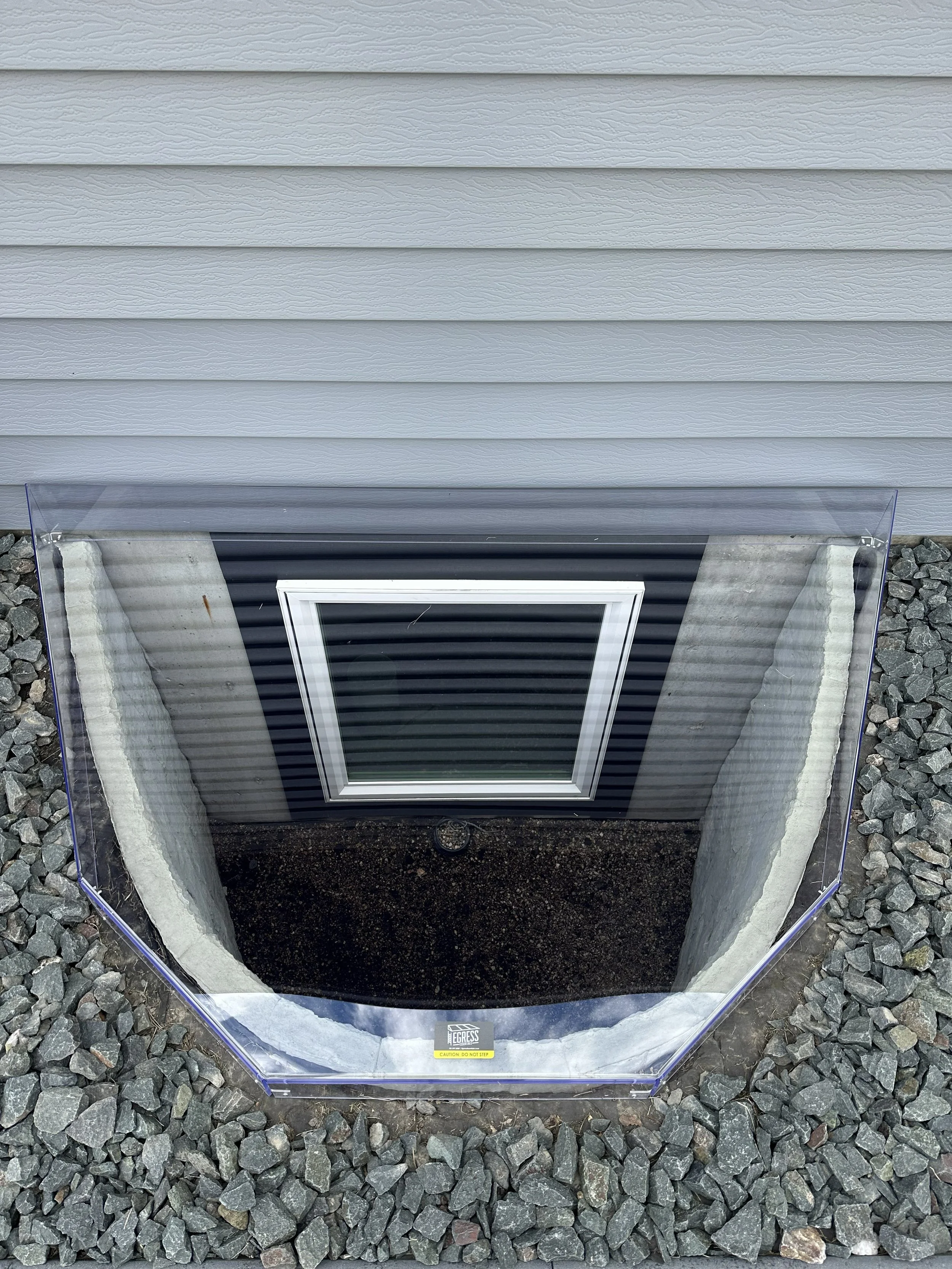 A rectangular grind opening in the ground with a protective transparent cover, surrounded by gray rocks, beneath a house with gray vinyl siding.