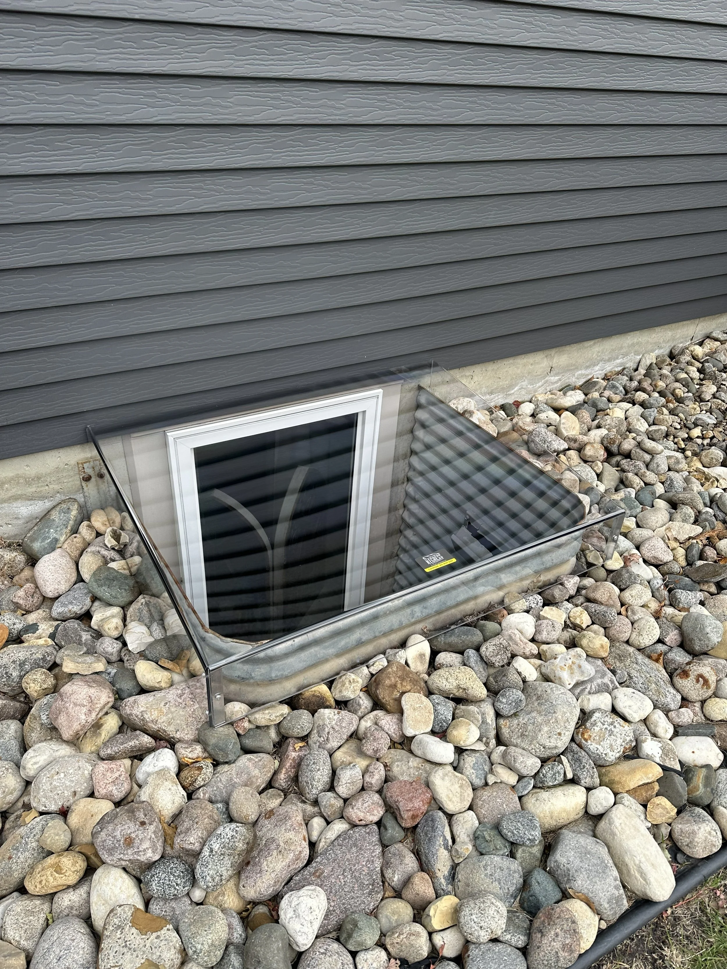 A basement window with the glass removed, metal framing, and a screen, set into a cement foundation wall. The area around the window is bordered by multicolored rocks.