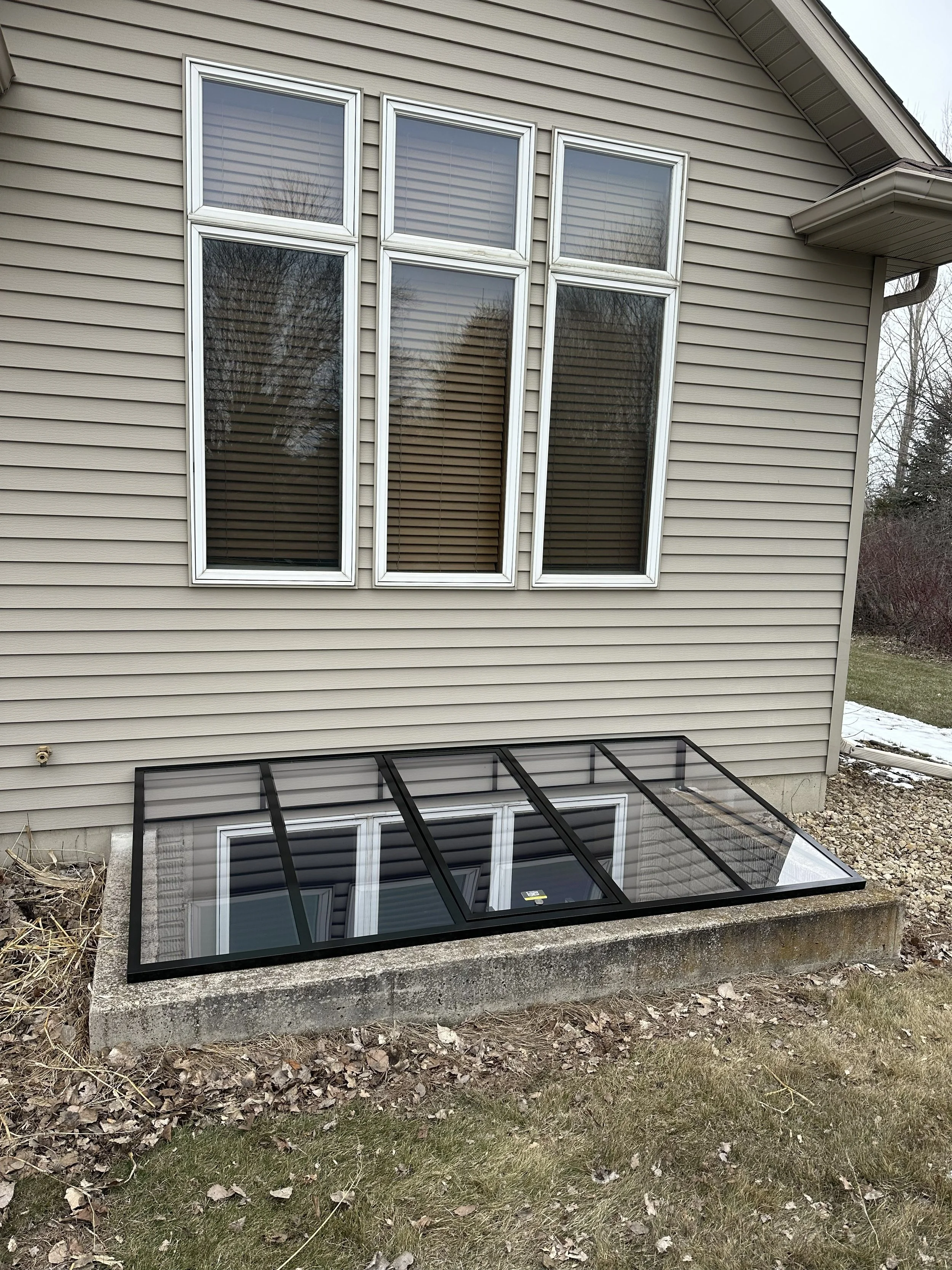 A house with beige siding and six large windows, with a small black-framed glass rooftop window at the bottom center, sitting on a concrete slab in the yard.