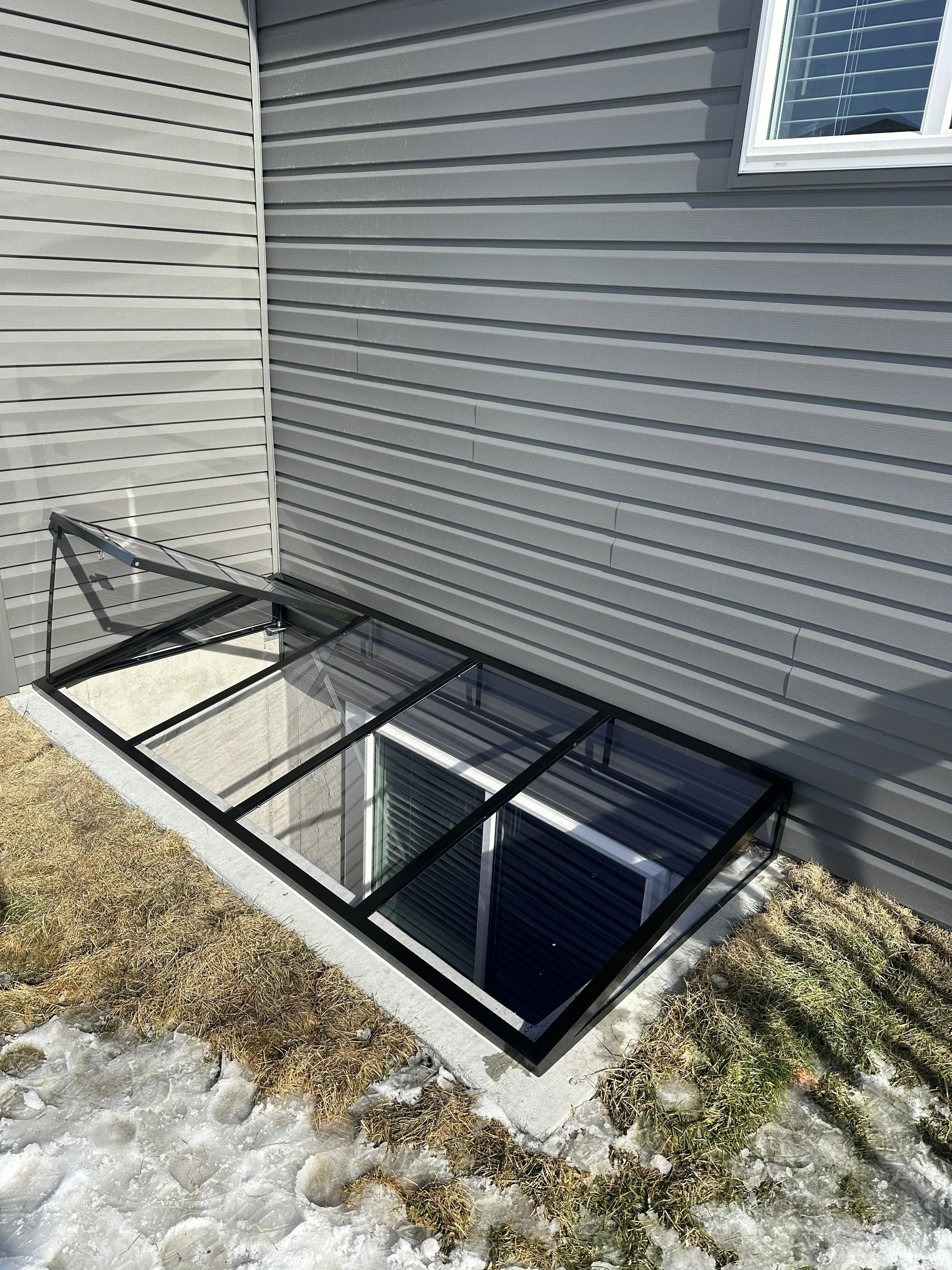Shed window with a glass roof and siding, with snow and patches of grass outside.