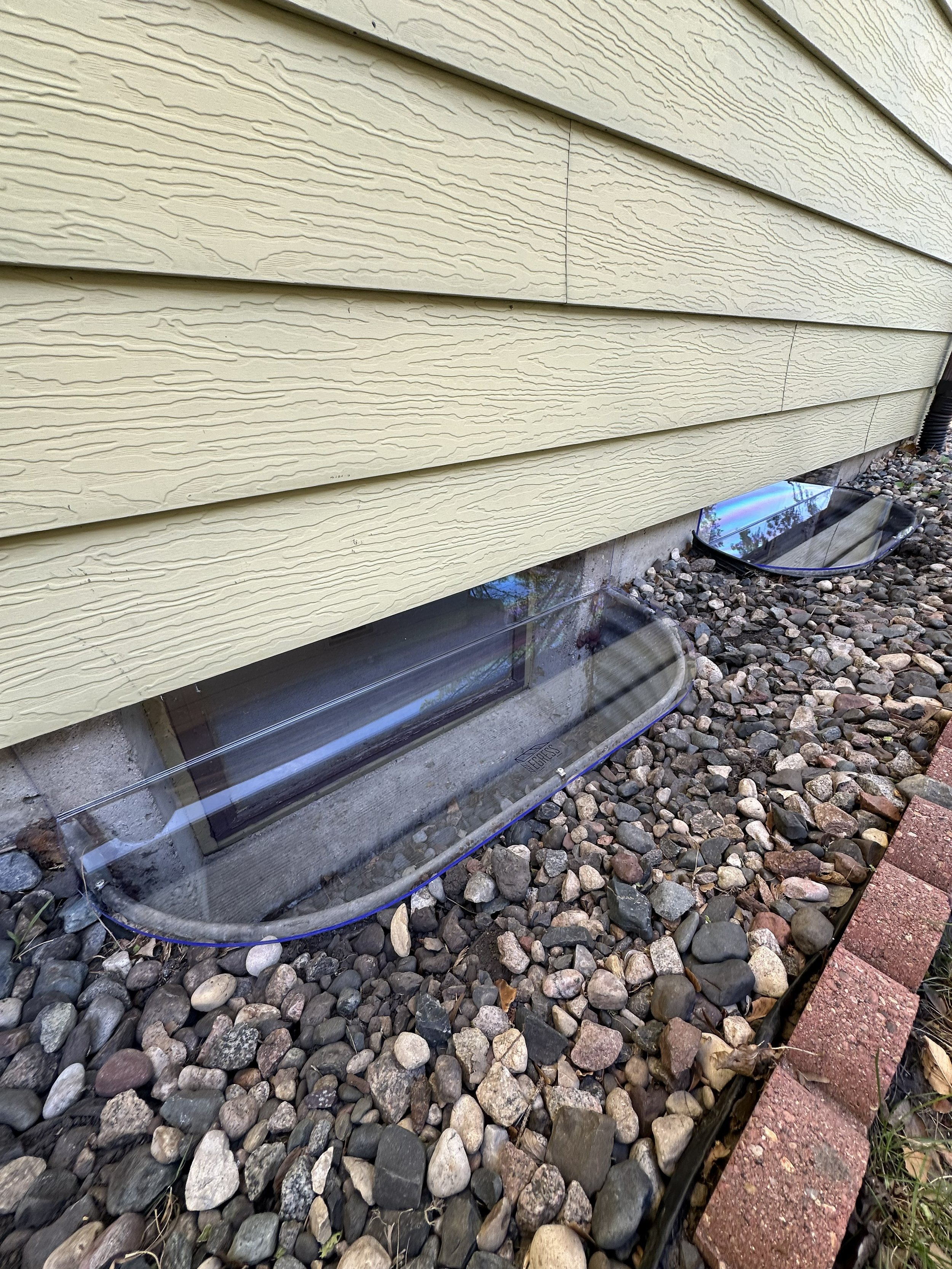 The image shows a close-up of the exterior siding of a house with beige textured panels. Beneath the house, there are two small, clear, rectangular window wells made of plastic or glass, with gravel surrounding them. Part of a brick pathway is visibl