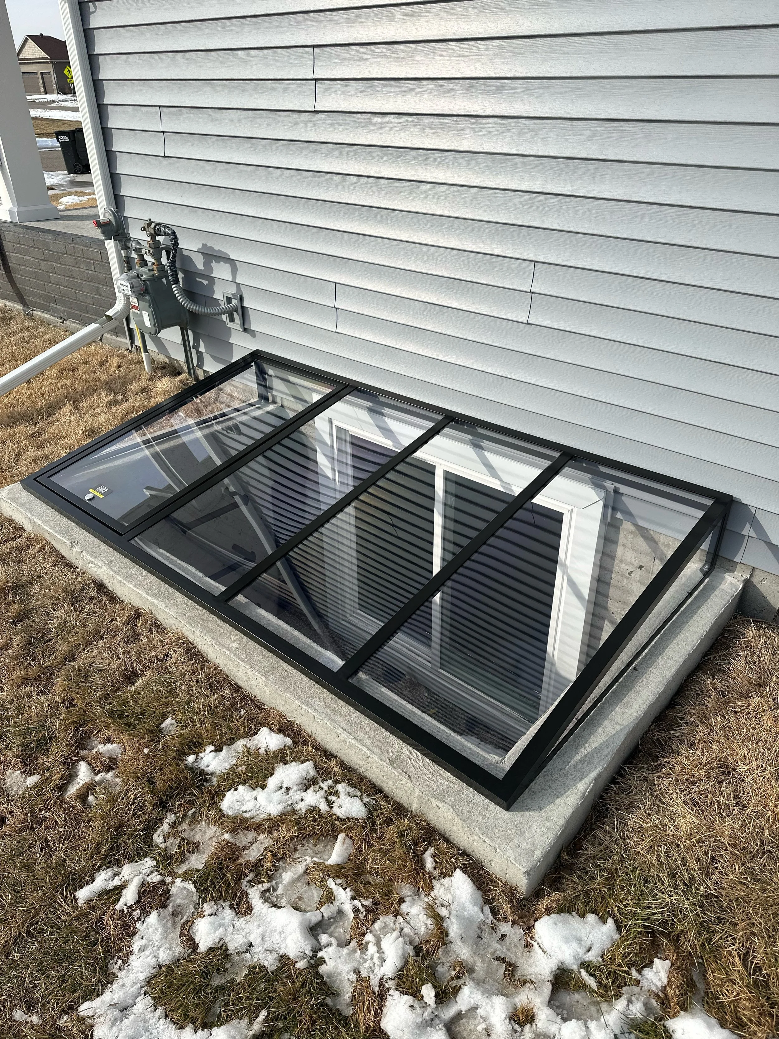 Photo of a house exterior with gray siding, a small basement window with a glass cover, and a gas meter with pipes on the side. There is snow on the ground and patches of dead grass.
