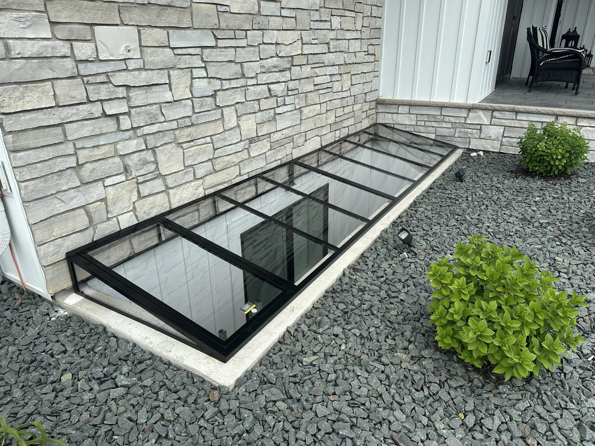 A backyard with a stone wall, gravel ground, a small shrub, and a glass-covered underground vent or window