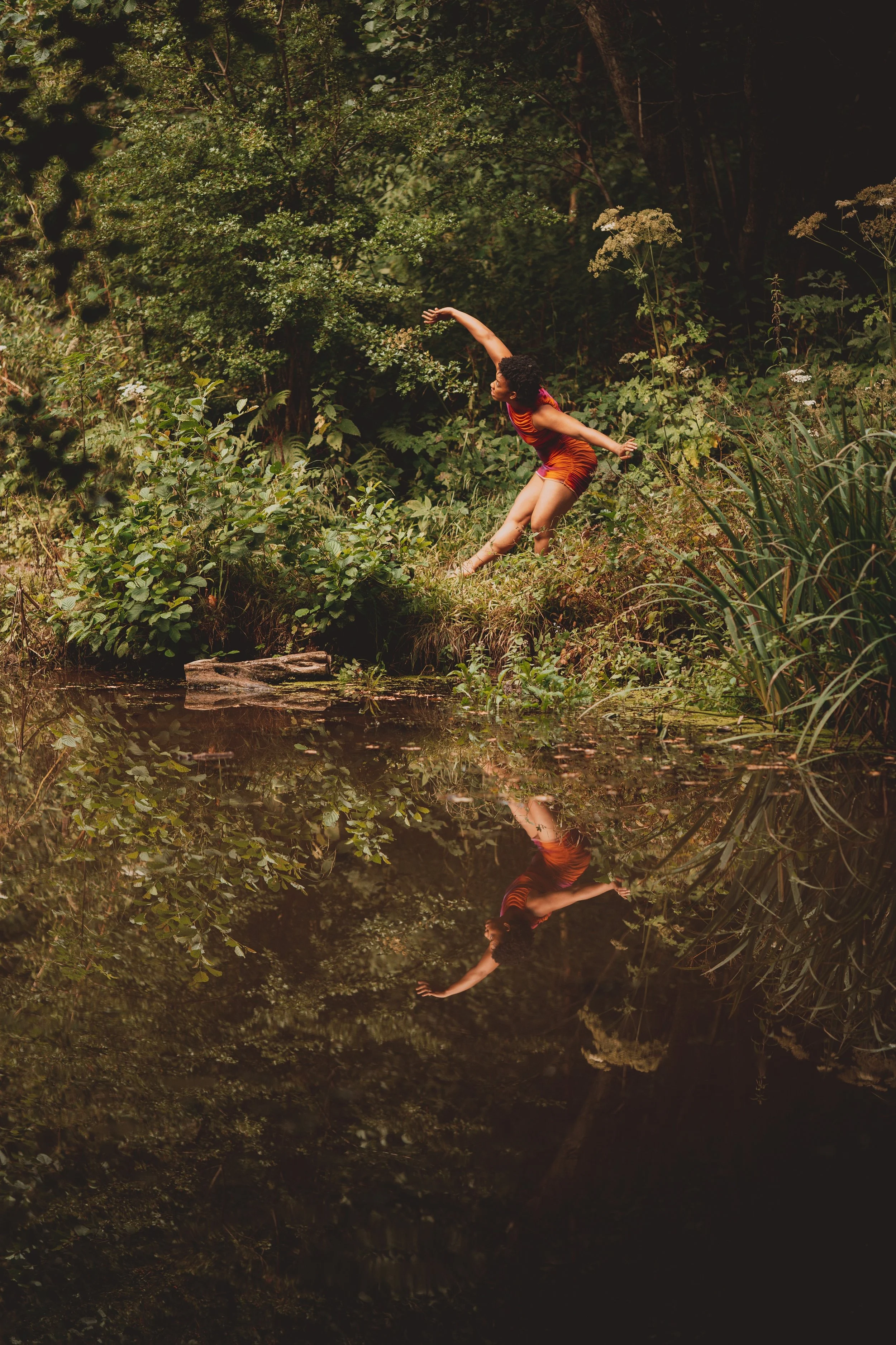 She stands seemingly engulfed by the forest around her and between you and her, there rests a lake of water. She bends slightly forward as her leg reaches outward and her right arm reaches to the trees behind her.