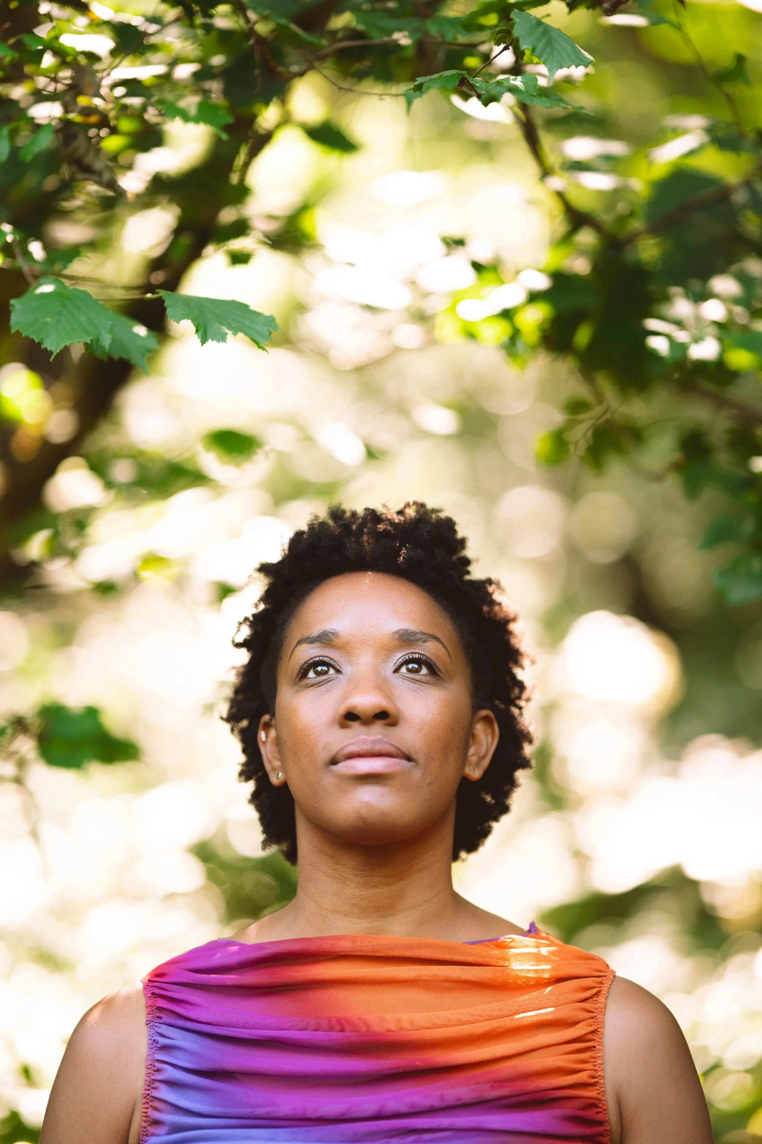 Headshot Image: The woman stands directly facing you but her eyes look upward. Only her head and shoulders are seen and there is the subtle emergence of leaves blurred behind her and the sun shines brightly.