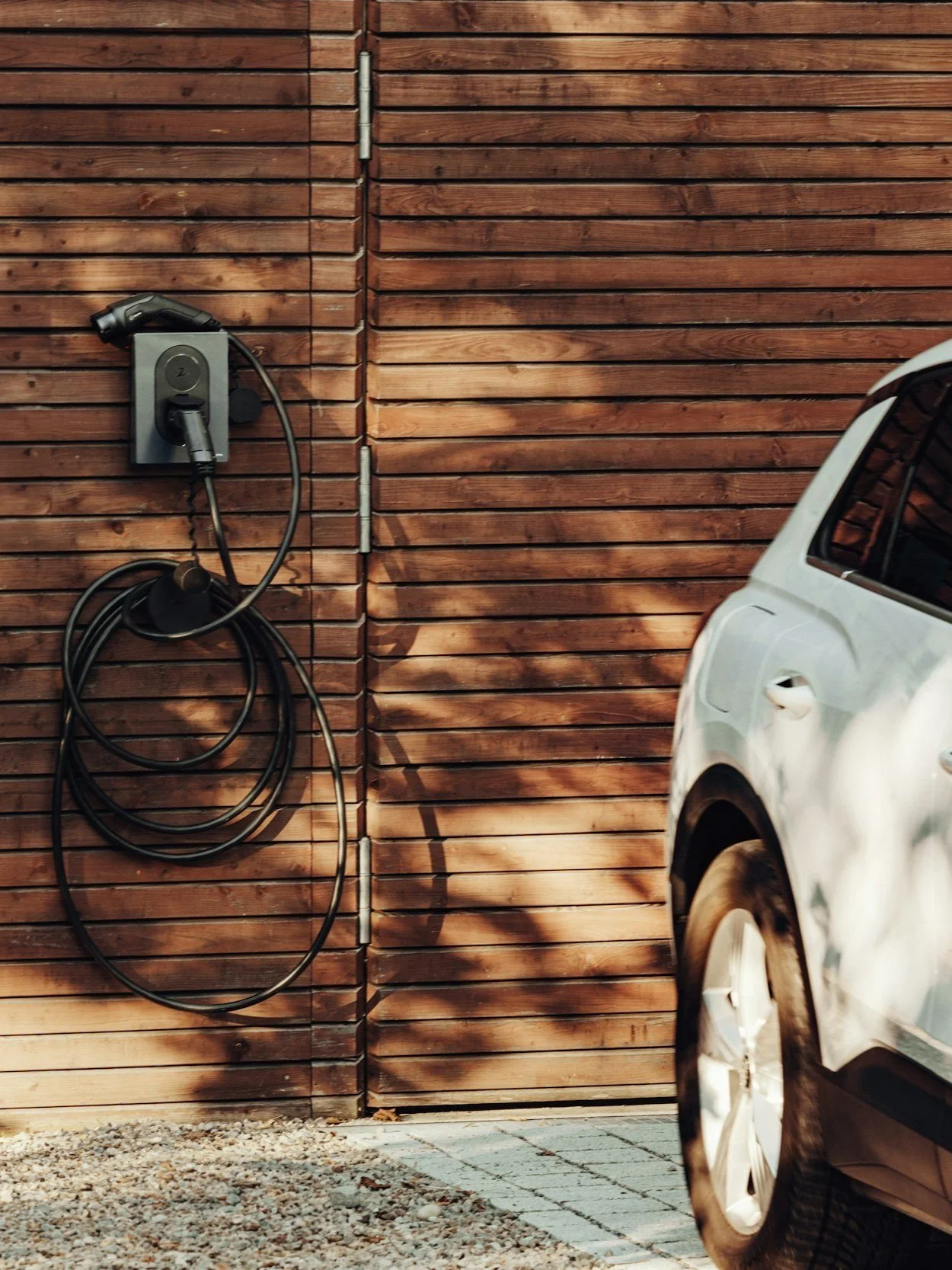A white electric vehicle charging station installed on a wooden fence with an attached charging cable, next to part of a white electric car parked on a gravel driveway.