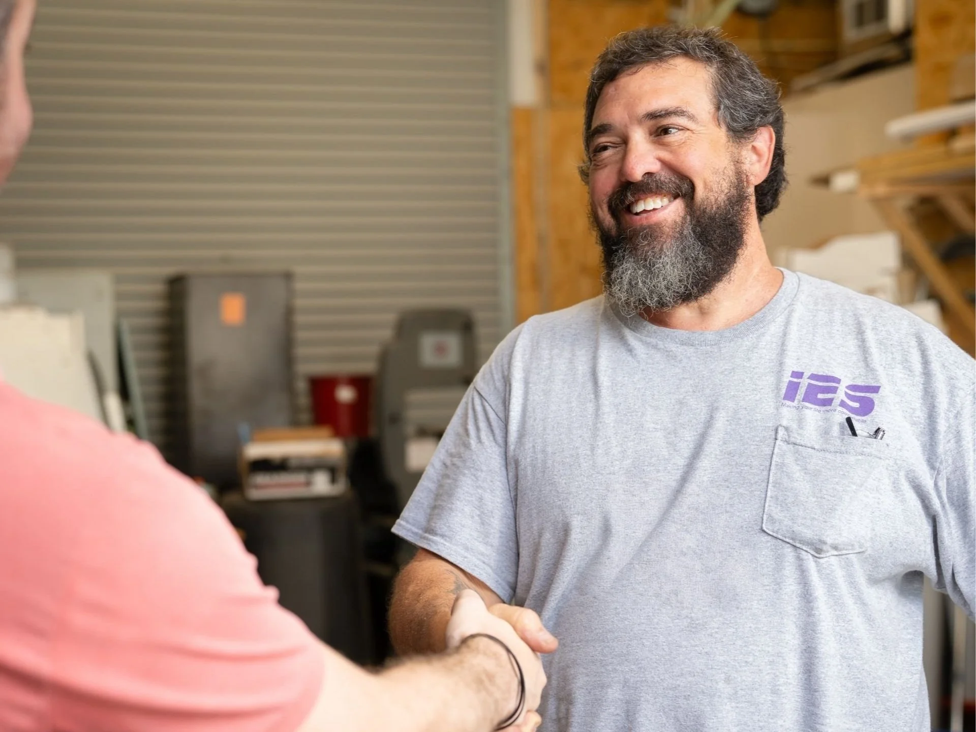 A man with a beard smiling while shaking hands with another person in a workshop or warehouse setting.