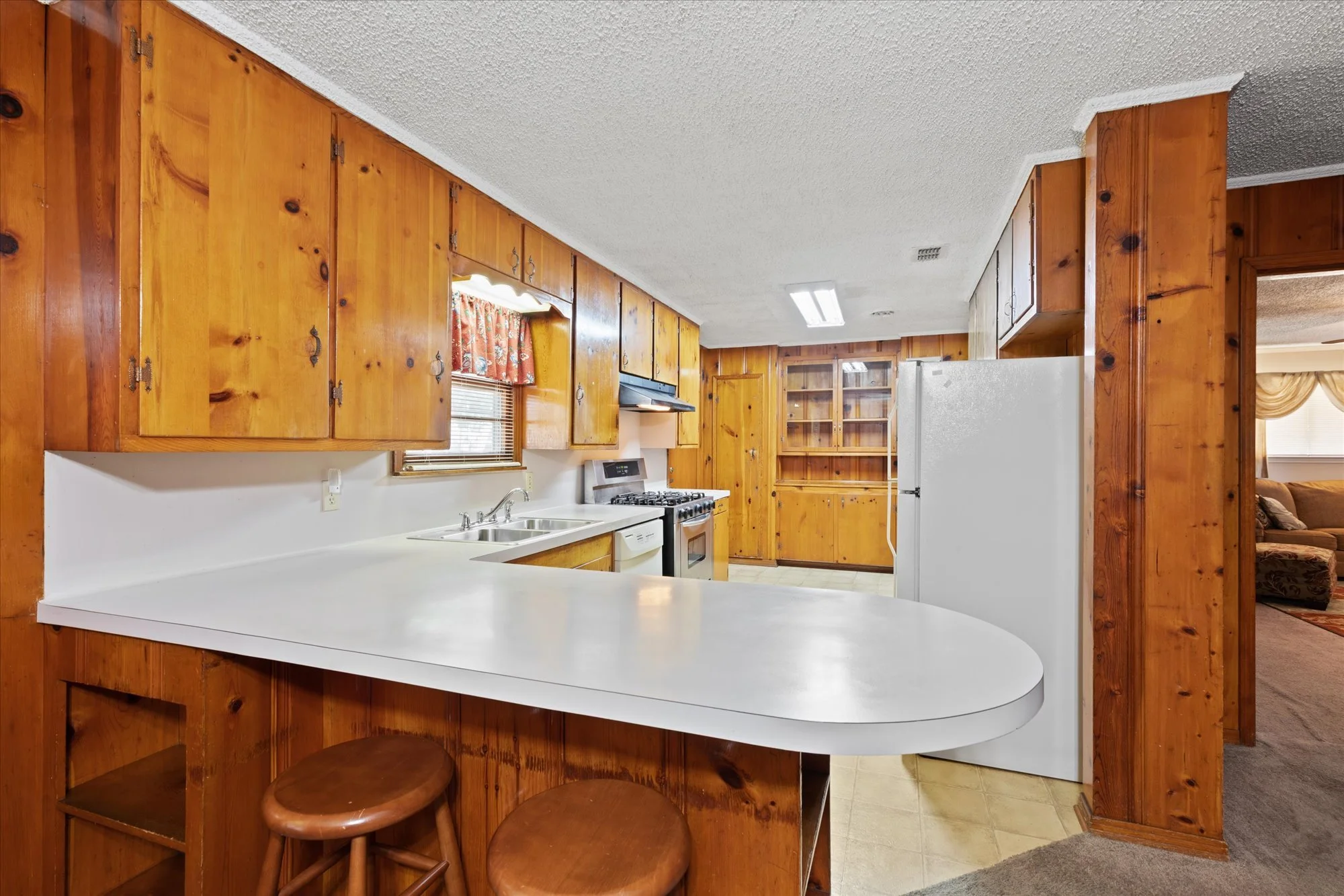 Wood-paneled kitchen with white countertops, stove, sink, refrigerator, and two wooden stools at the breakfast bar.