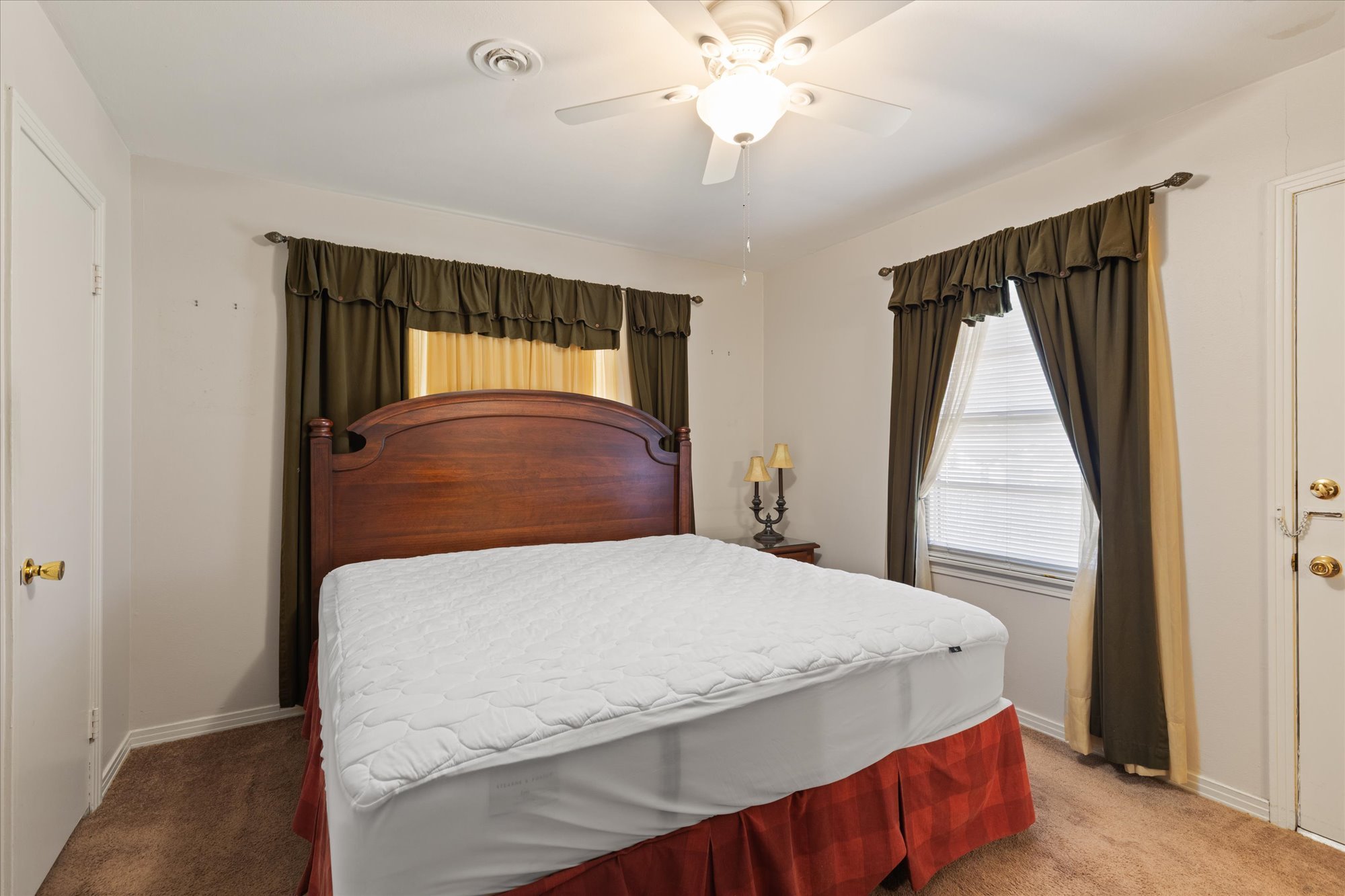 Empty bedroom with a large bed, wooden headboard, white mattress, two curtains on windows, wall lamp, ceiling fan, carpeted floor, and plain walls.