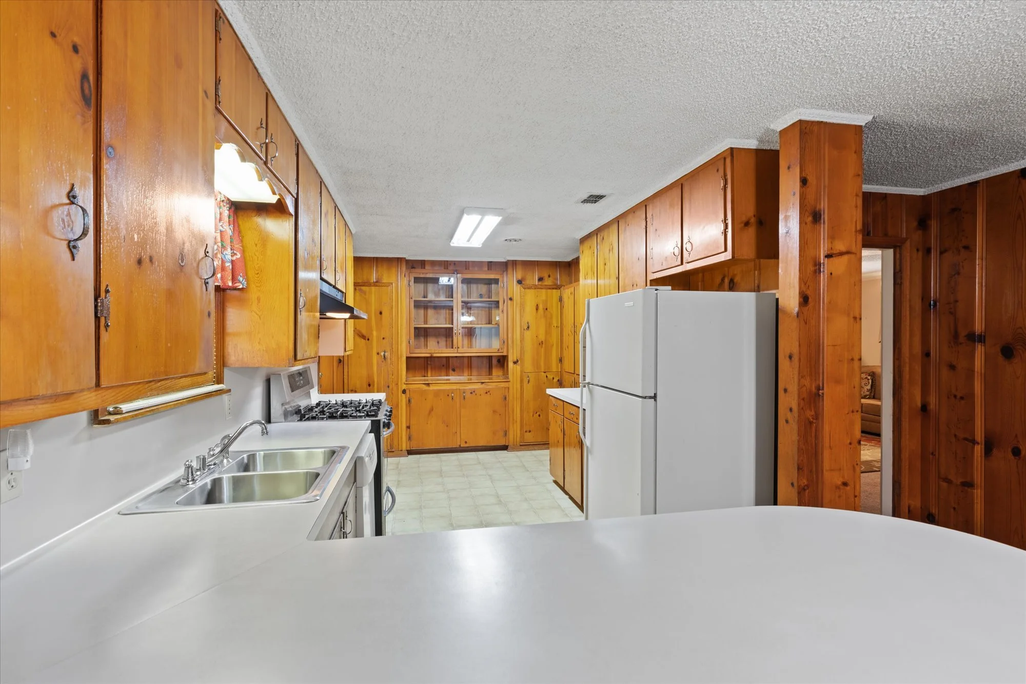 View of a kitchen with wooden cabinets, white refrigerator, gas stove, and stainless steel sink, with a doorway leading to a living room.