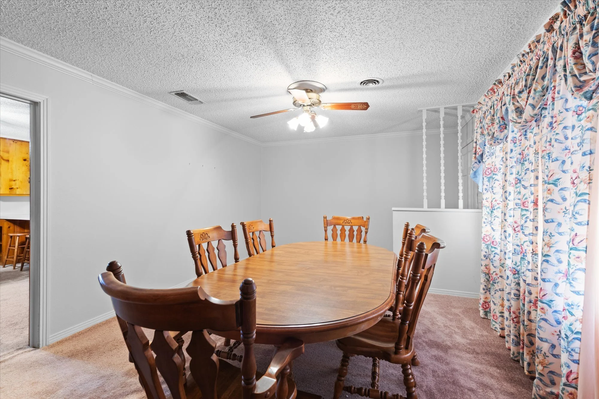 Dining room with wooden table and chairs, floral curtains, white walls, ceiling fan with lights, beige carpeted floor.