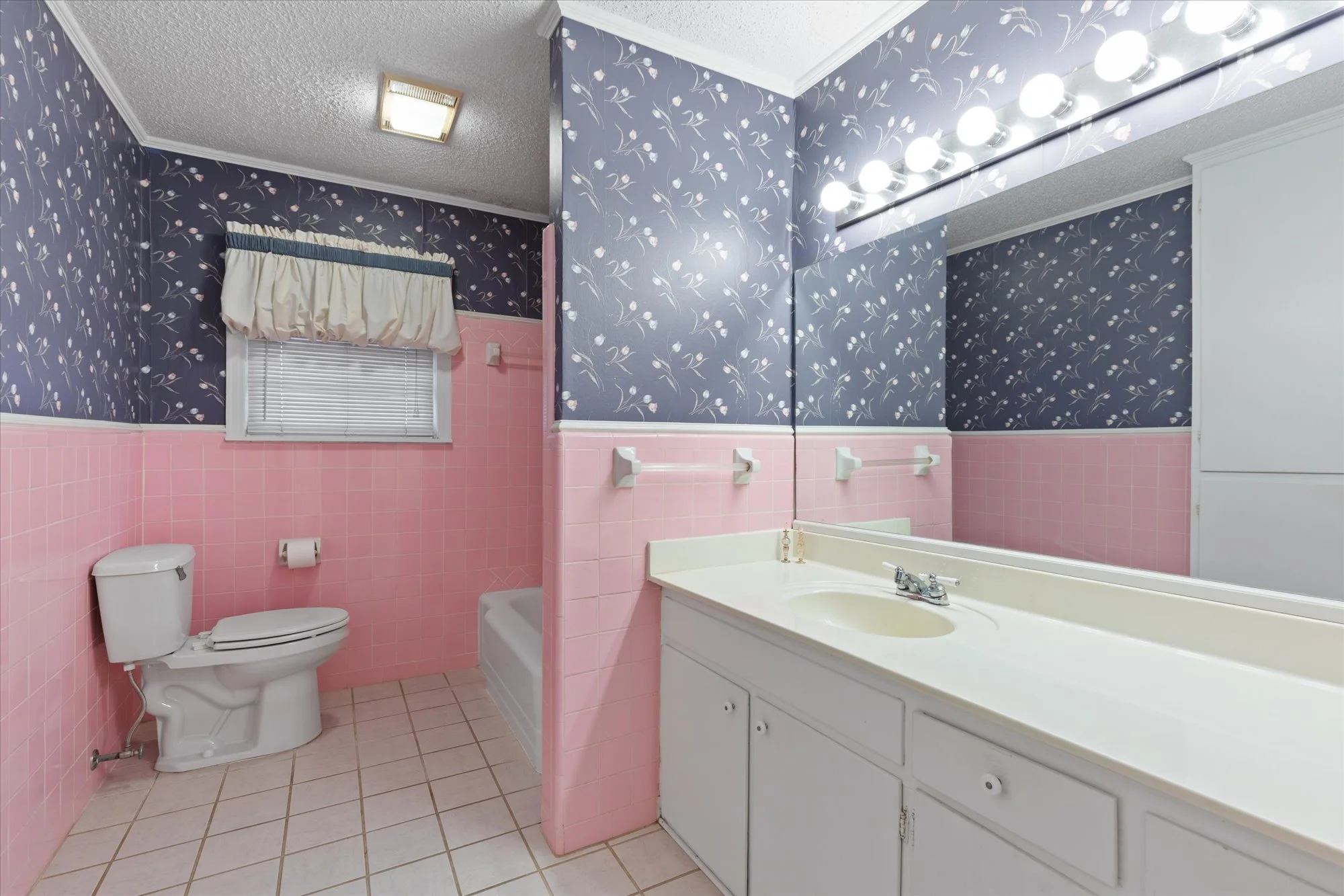 A bathroom with pink tiled walls halfway up and dark blue walled wallpaper with floral pattern on the upper part, a white toilet, a window with a beige valance, a bathtub, and a sink with a large mirror and bright lights.