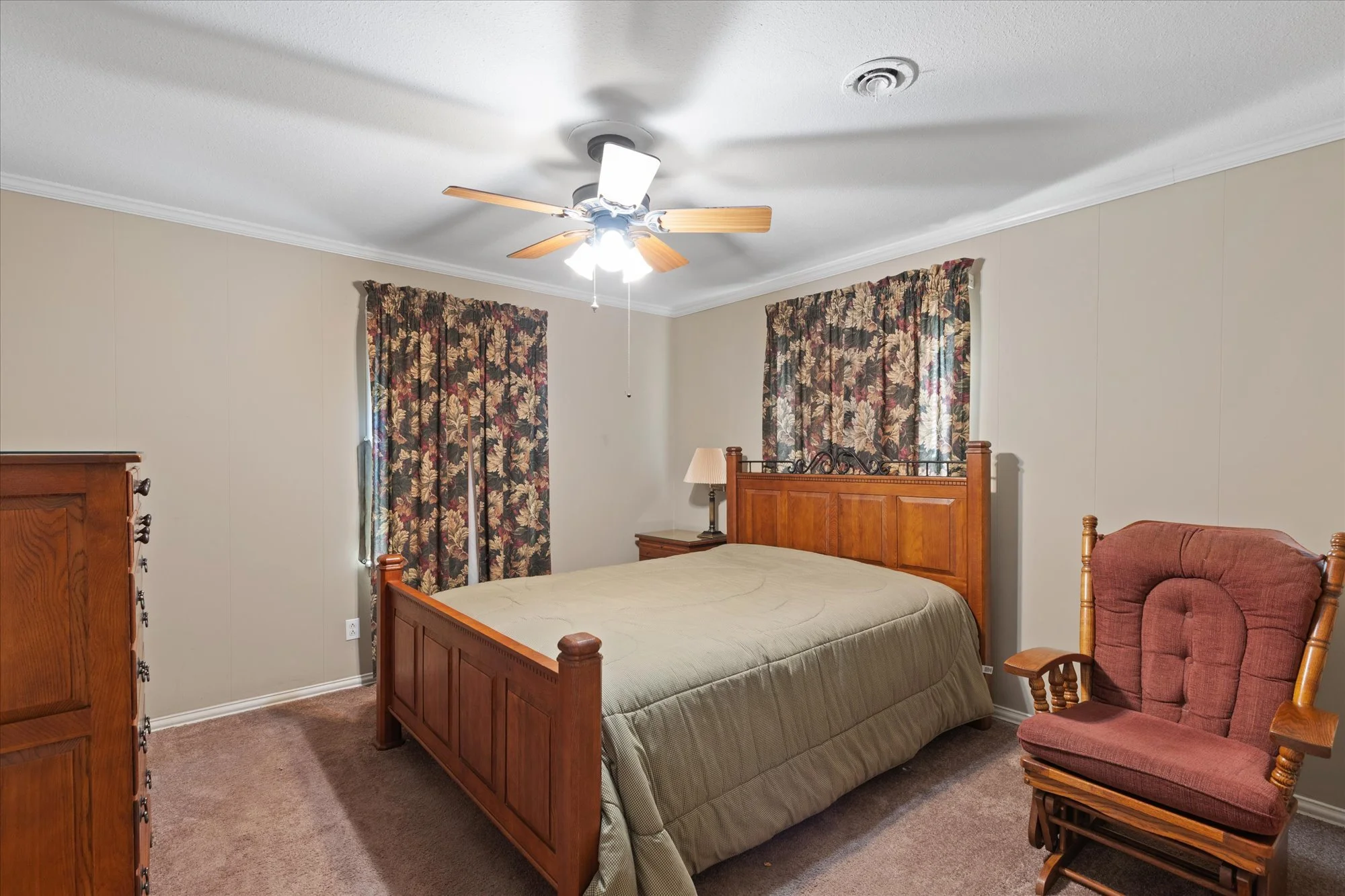 Bedroom with wooden bed frame, beige bedspread, floral curtains, wooden dresser, antique wooden rocking chair, and beige walls with white crown molding