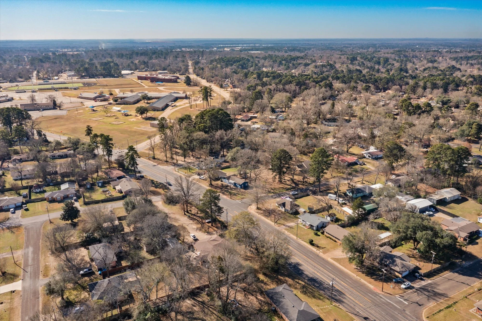 Aerial view of a residential neighborhood with houses, trees, and streets, adjacent to a school campus with athletic fields and buildings, under a clear blue sky.
