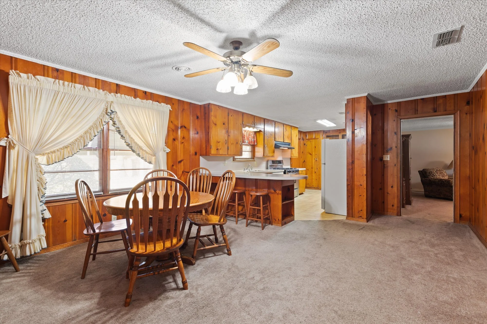 A cozy dining area and kitchen with wood-paneled walls, a window with cream-colored ruffled curtains, a round wooden dining table with five matching chairs, a ceiling fan with wooden blades, a kitchen counter with stools, a stove, a refrigerator, and