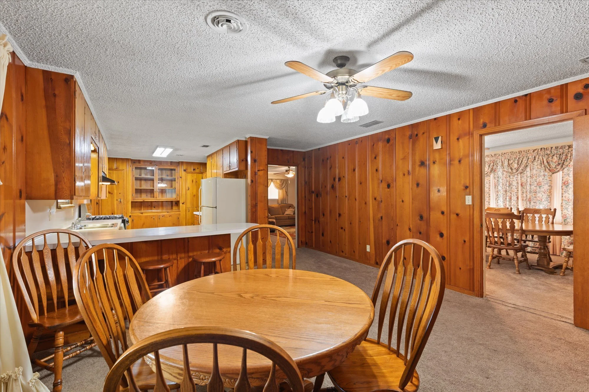 Wood-paneled dining area with a round wooden table and six matching chairs, leading to a kitchen with a white refrigerator and open cabinets, and into a living room with curtains.
