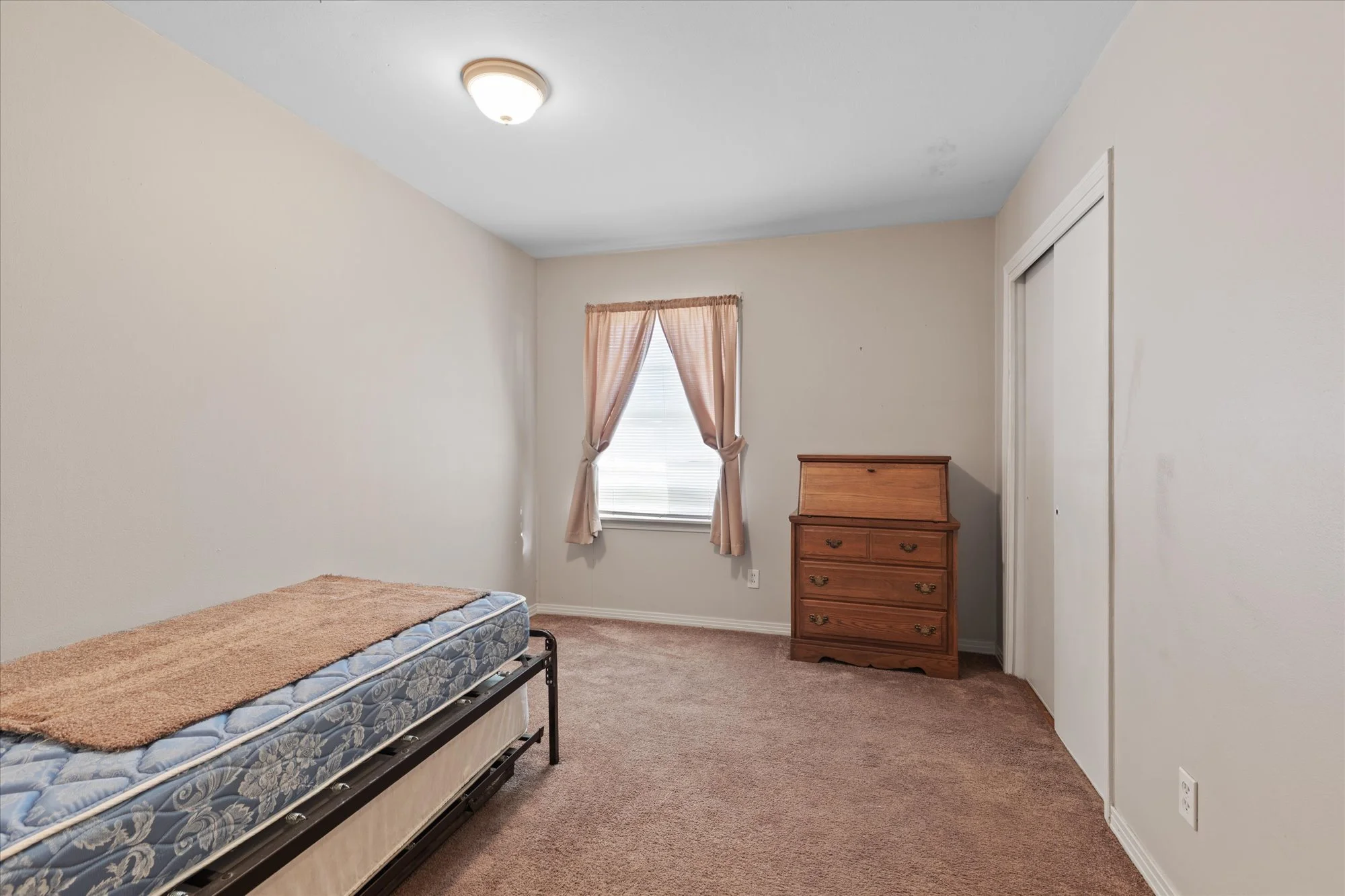 Empty bedroom with beige walls, a window with pink curtains, a wooden dresser, and a mattress on a metal frame.