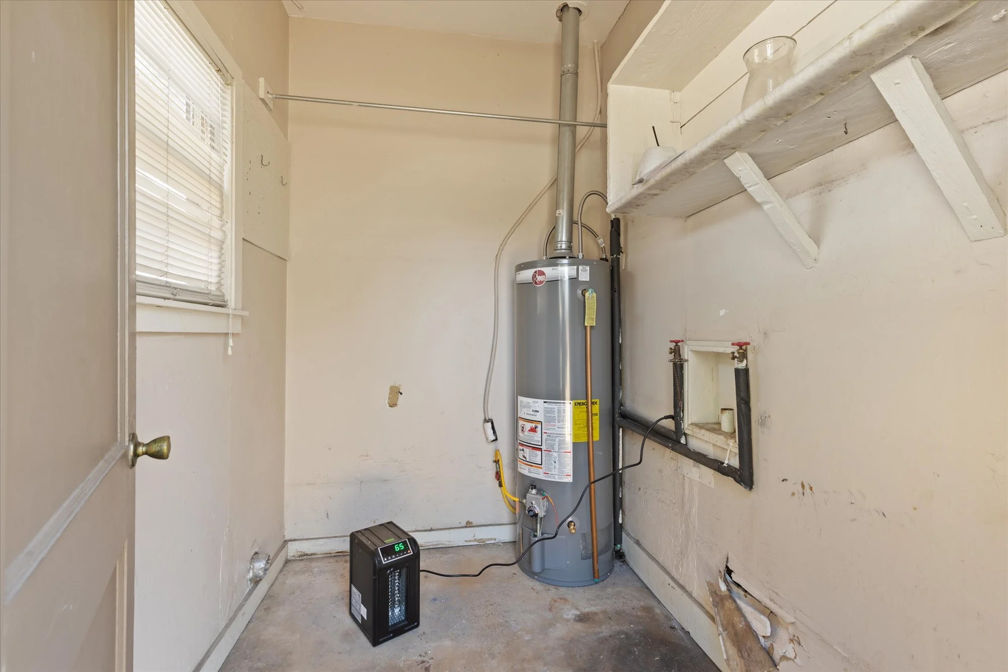 A water heater in a small laundry room with a heater on the floor displaying 65 degrees, and a window with closed blinds.