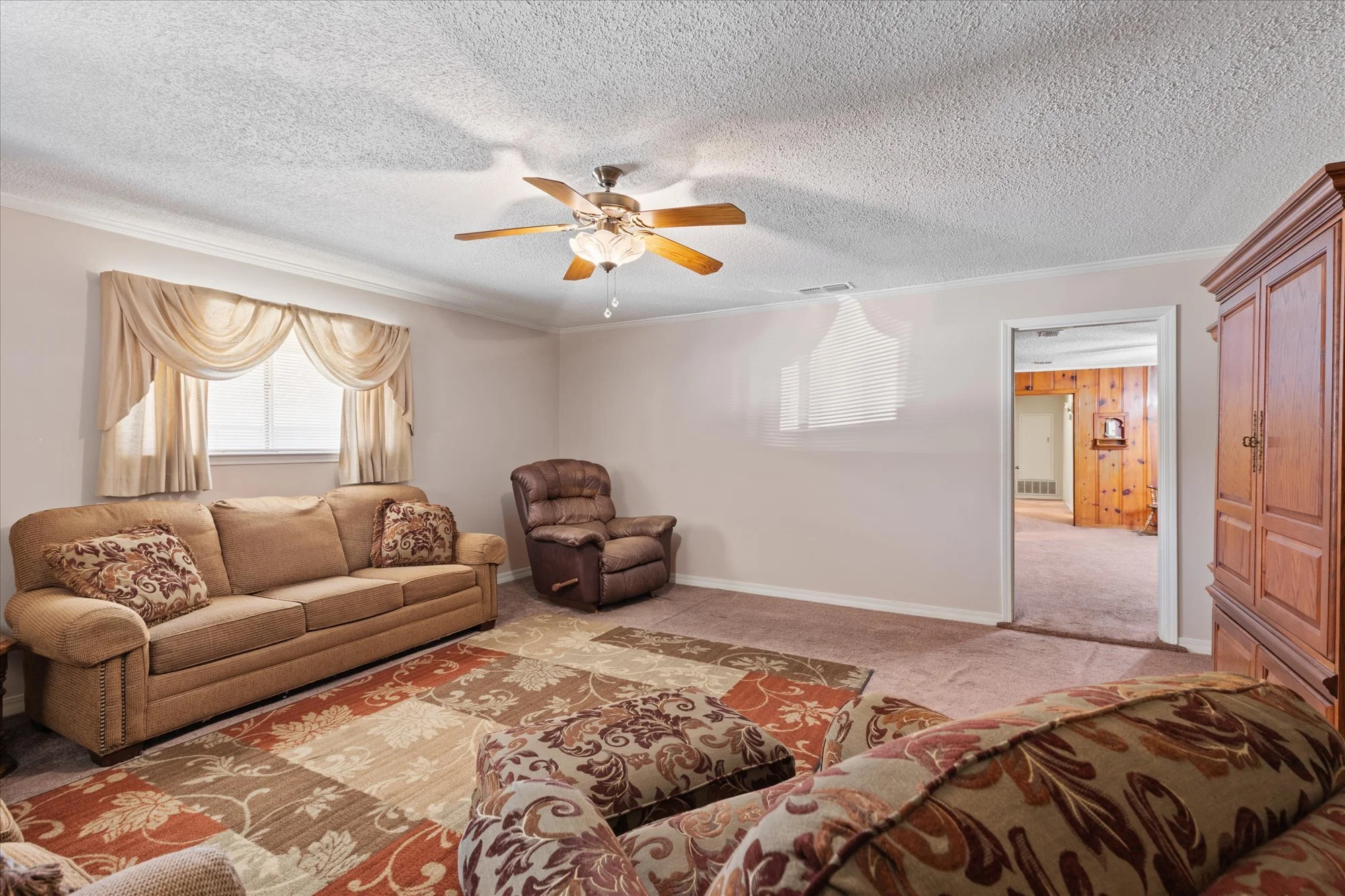 Living room with beige sofa, patterned pillows, patterned area rug, ceiling fan, window with beige drapes, brown recliner, and wooden furniture, with an open doorway leading to another room.