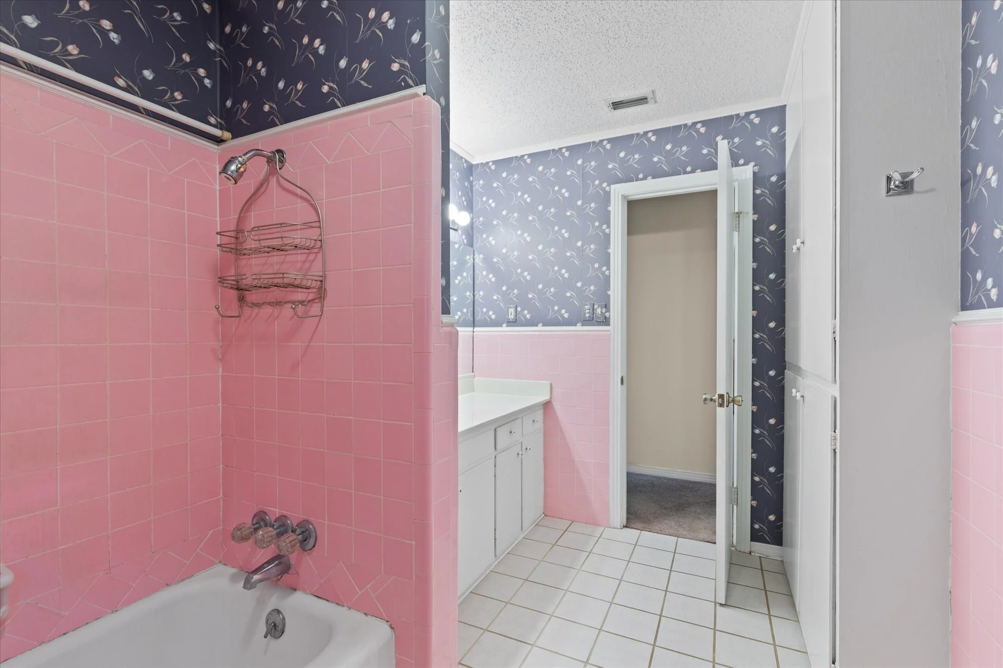 Bathroom with pink tiled bathtub area, navy blue wallpaper with white floral pattern, white vanity with mirror, and an open door leading to a linen closet.