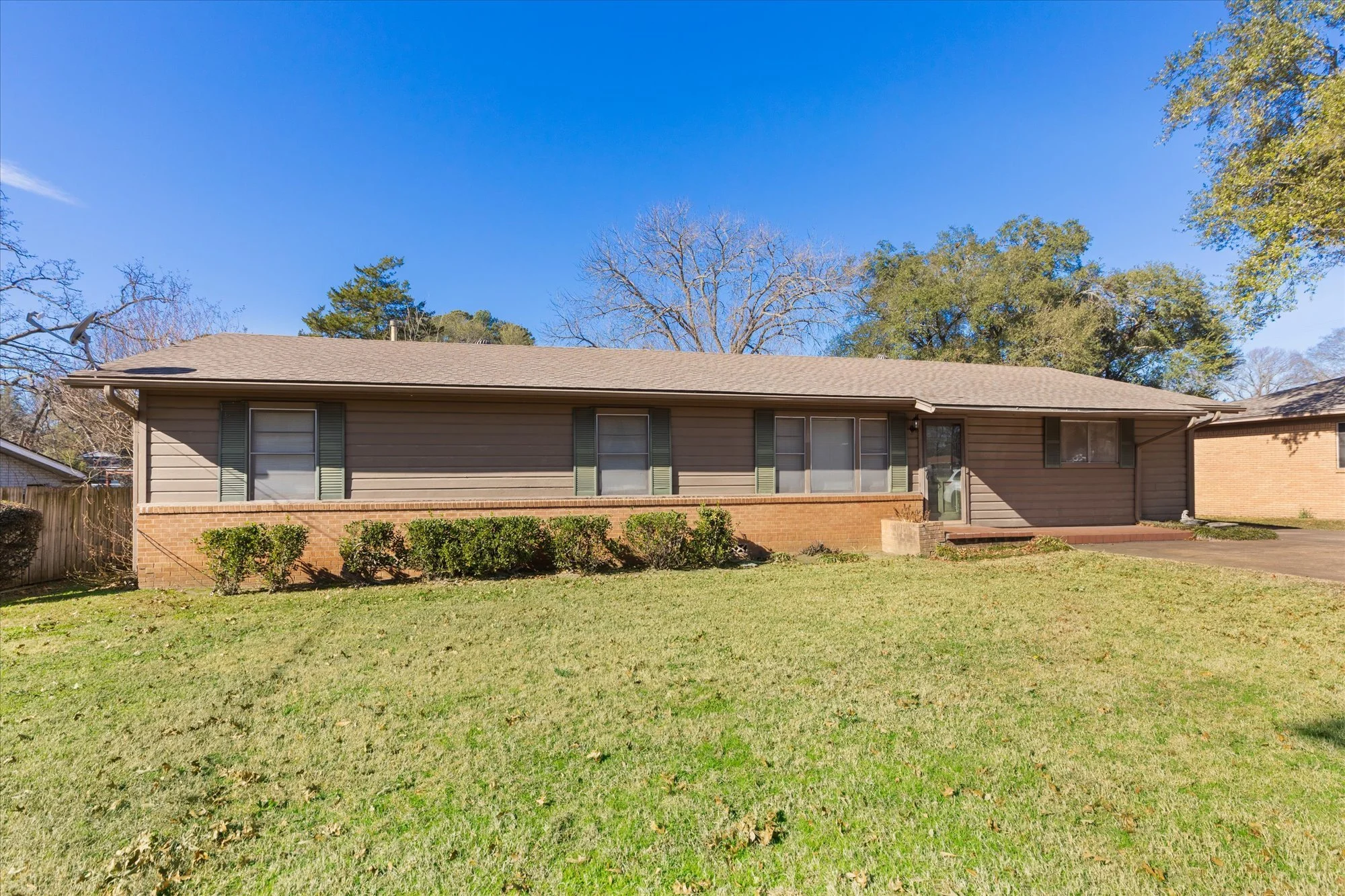 A single-story house with beige siding, green shutters, and a brick foundation, surrounded by a lawn with small bushes, under a clear blue sky.