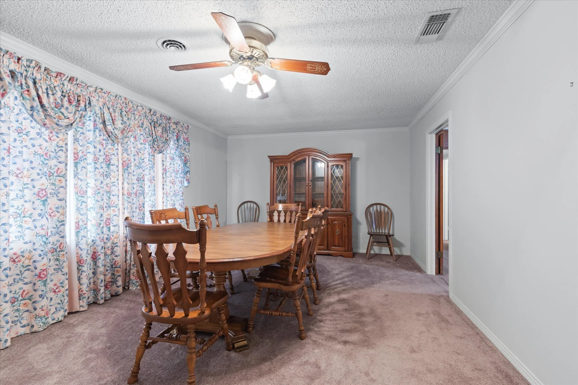 Dining room with a wooden table and six matching chairs, a wooden china cabinet, a chair near the wall, floral curtains over windows, white walls, beige carpet, ceiling fan with light, and an open doorway.