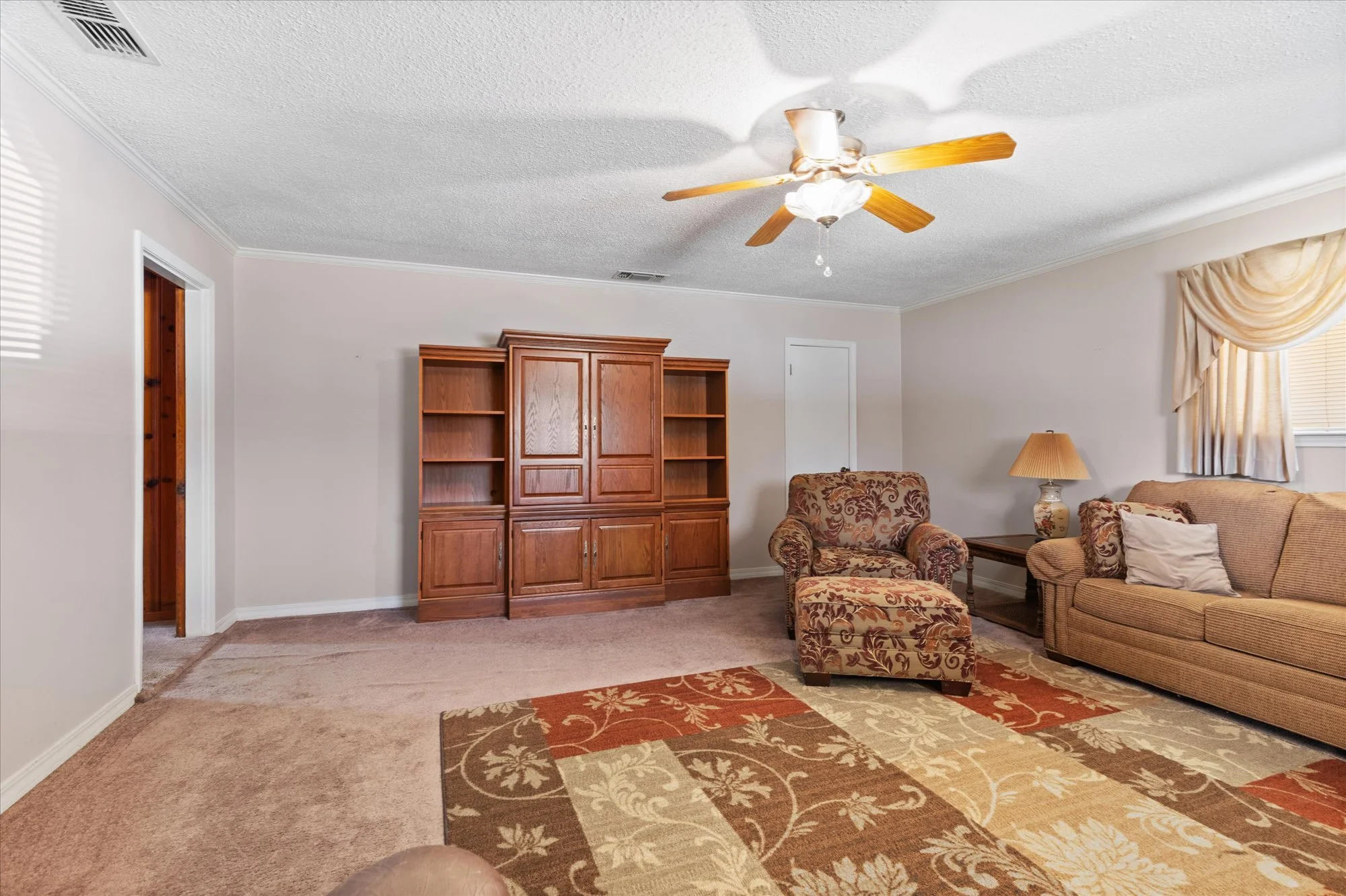 Living room with beige carpet, a wooden entertainment center, a floral armchair with matching ottoman, a beige sofa with pillows, a side table with a lamp, a window with curtains, and a ceiling fan.