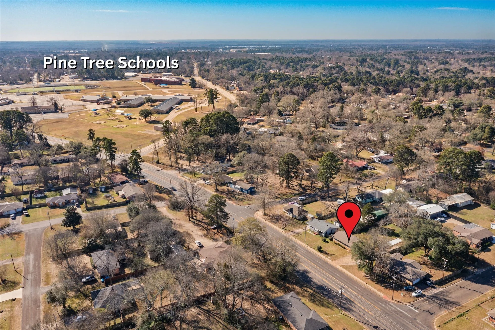 Aerial view of a neighborhood with houses and trees, a main road running through, and Pine Tree Schools in the background, with a red location pin indicating a specific house.
