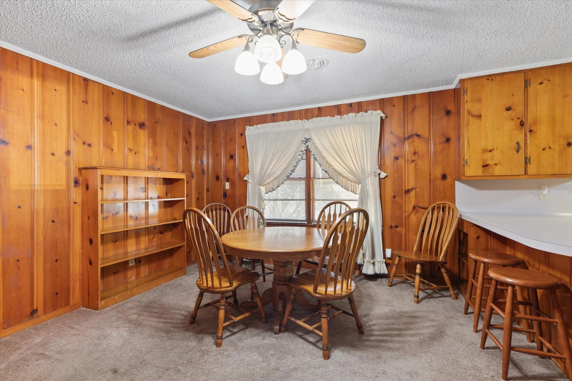 A dining room with wood-paneled walls, a round wooden table with six matching chairs, a window with white curtains, a ceiling fan with lights, a small built-in bookshelf, and a kitchen counter with stools.