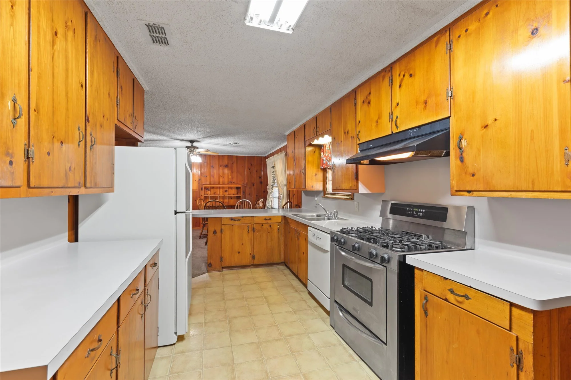 A kitchen with wooden cabinets, white countertops, a white refrigerator, a dishwasher, a stainless steel oven with a stove, a black range hood, a double sink under a window, and a wooden dining area in the background.