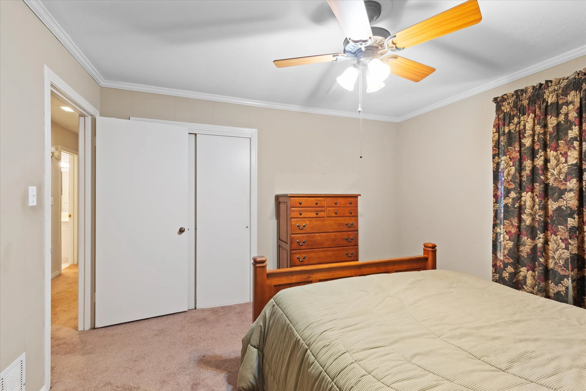 A bedroom with beige walls, a ceiling fan with wooden blades, a wooden dresser, a bed with a beige bedspread, and floral curtains.