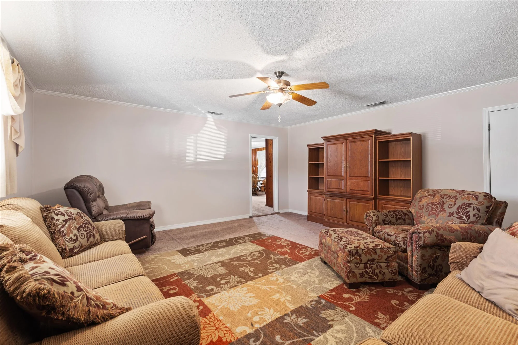 Living room with beige and floral-patterned sofas, a wooden entertainment center, a recliner, and a ceiling fan. A colorful area rug covers part of the carpeted floor. A doorway leads to a dining area.