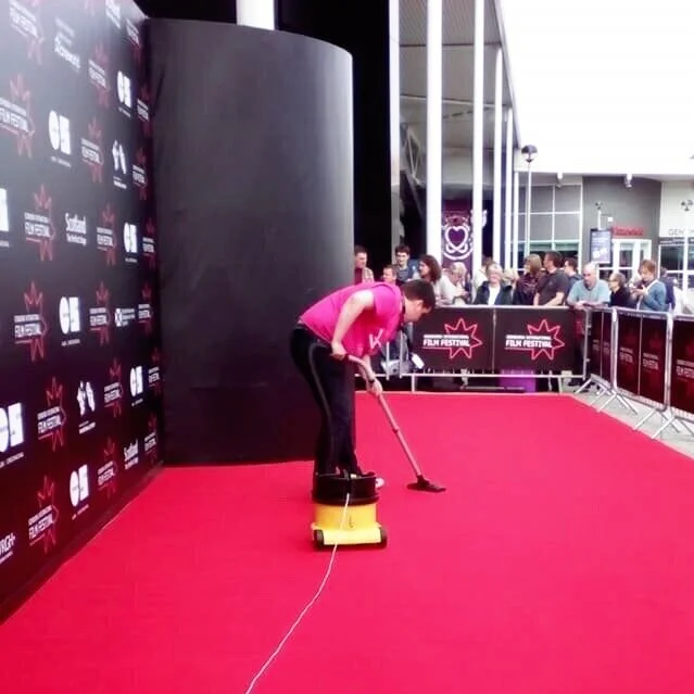 Person in a pink shirt cleaning a red carpet at an event with audience and barriers in the background.