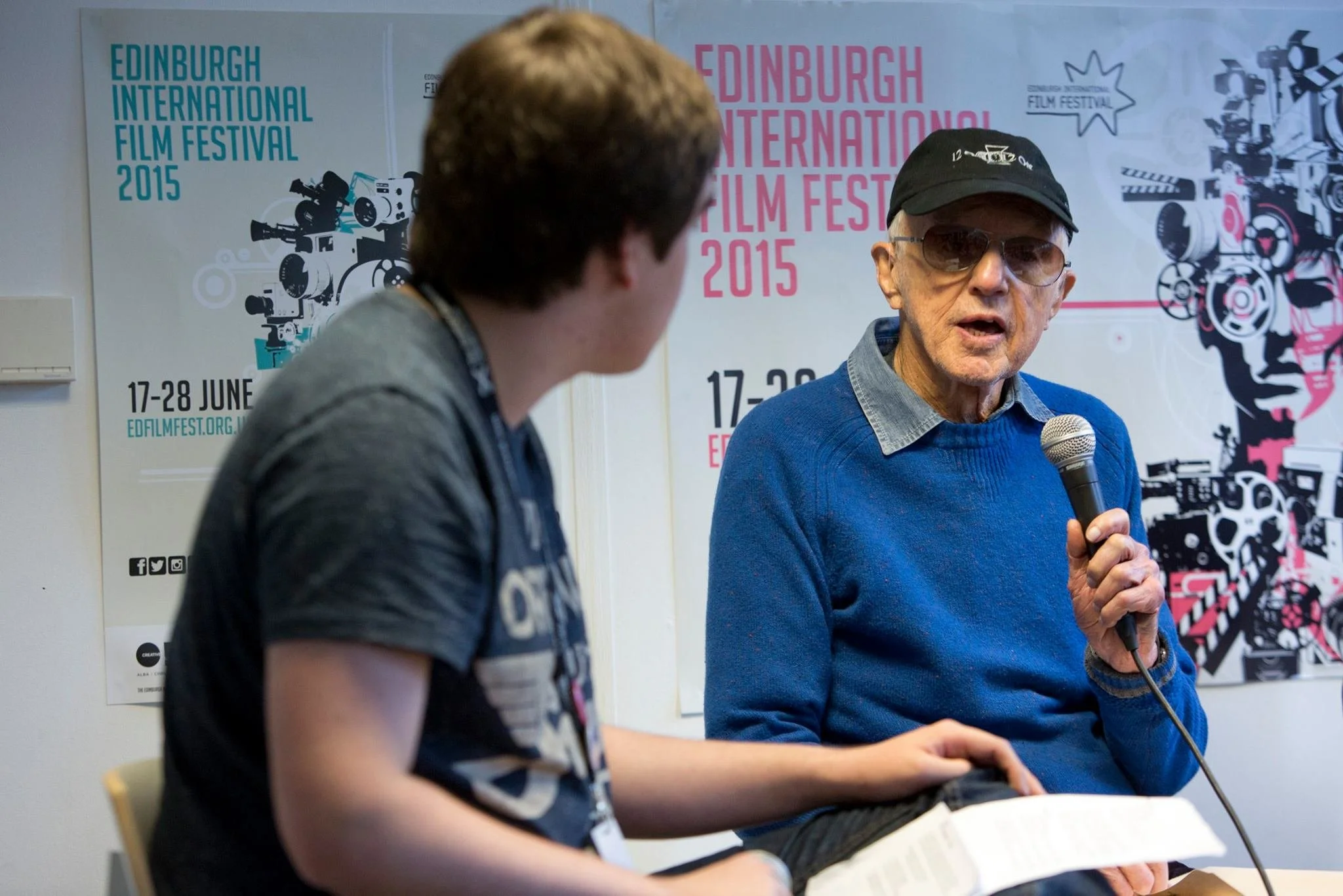 An elderly man wearing sunglasses and a black cap, holding a microphone, speaking during the Edinburgh International Film Festival 2015. A young person in a gray shirt is sitting next to him, looking at a book. Posters for the festival are visible in the background.