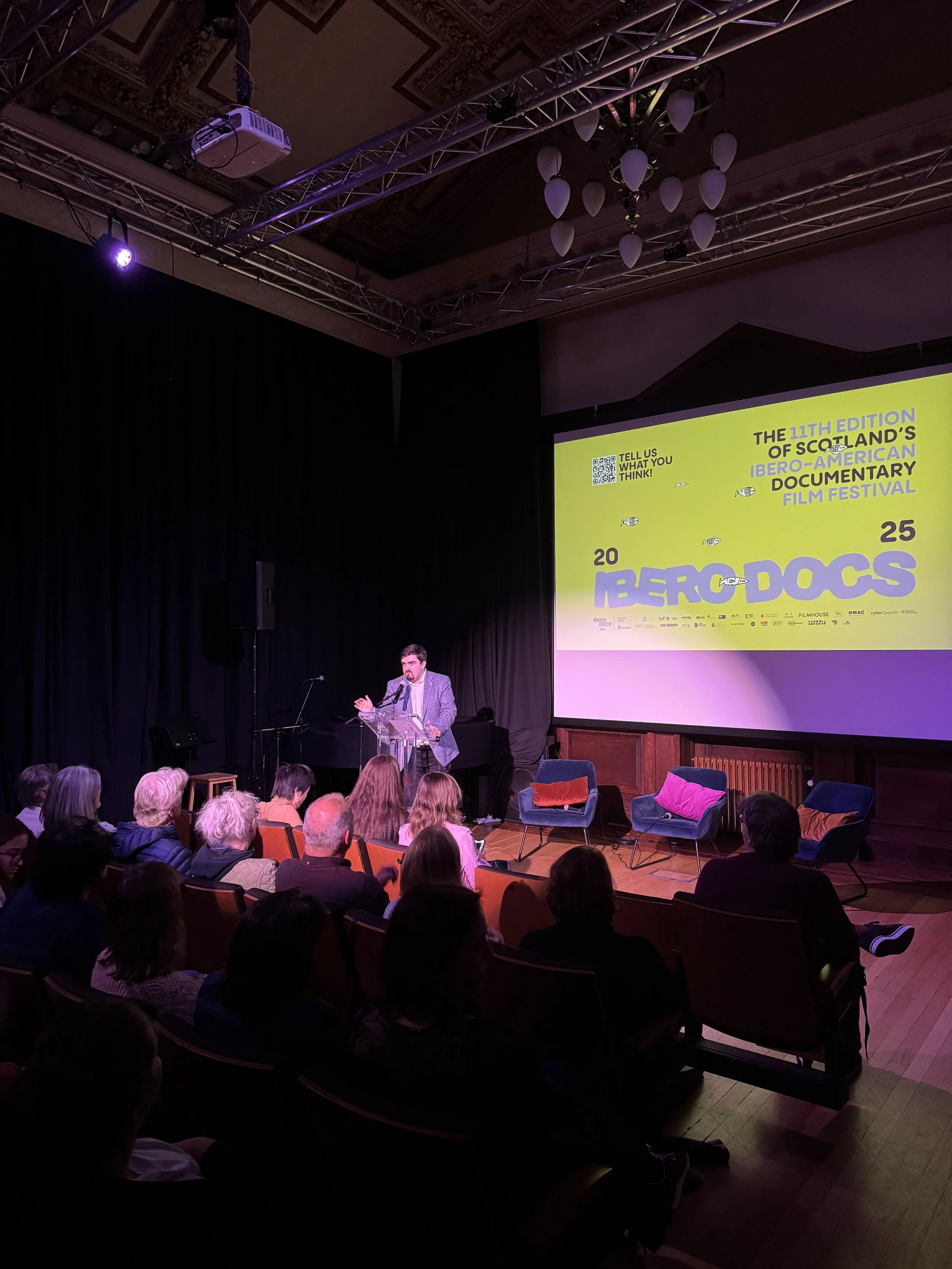 A man giving a speech at a podium during the Ibero-American Documentary Film Festival event, with a large yellow presentation slide behind him displaying event details and logos, audience seated facing him in a dimly lit indoor venue.