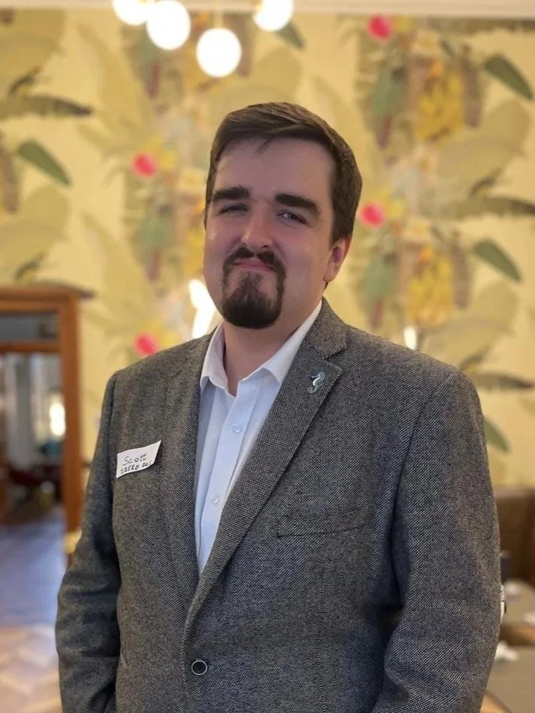 A man in a gray blazer and white shirt standing indoors with a floral wallpaper background and ceiling lights.
