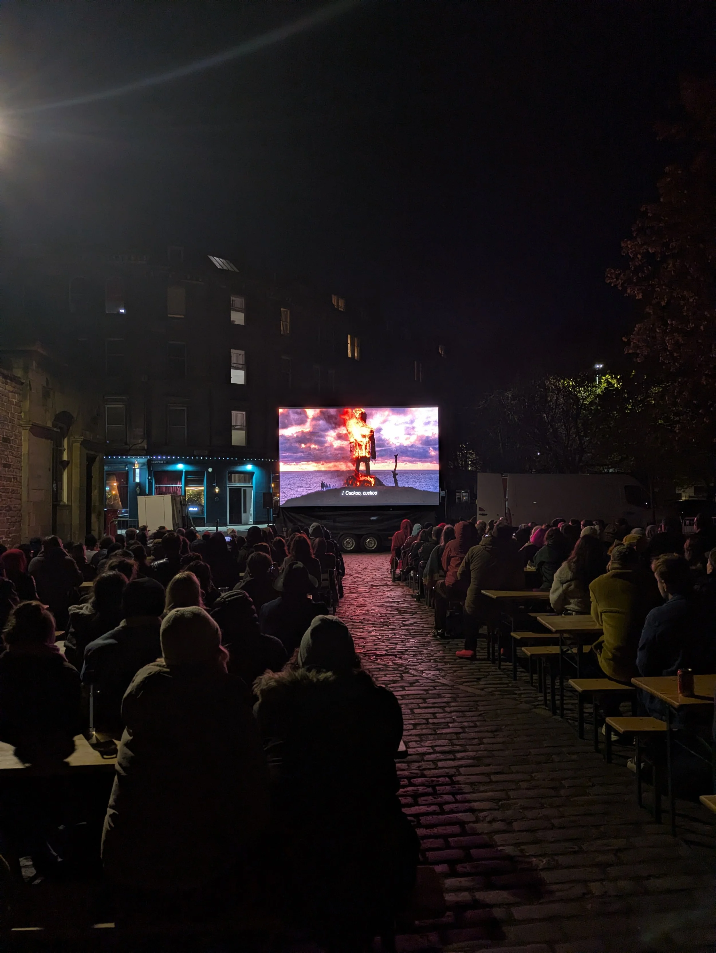 An outdoor movie theater at night with an audience sitting on benches and chairs, watching a movie on a large screen depicting a fiery scene near the ocean.