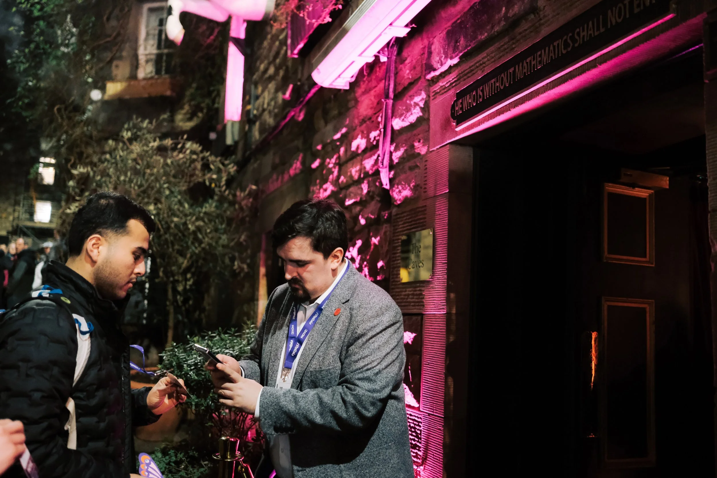 Two men stand in front of a brick building with pink lighting, one showing something on a phone to the other. Other people are visible in the background.