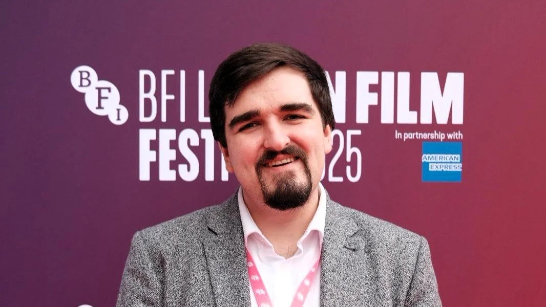 Man with dark hair, beard, and mustache wearing a gray blazer and pink shirt, standing in front of a purple background with the BFI London Film Festival logo and text.