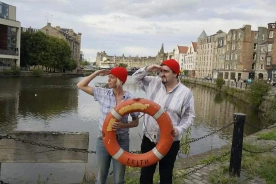 A woman and a man wearing red beanies and casual clothes, standing beside a river with historic buildings in the background, saluting and holding a life preserver labeled 'LEATH'.