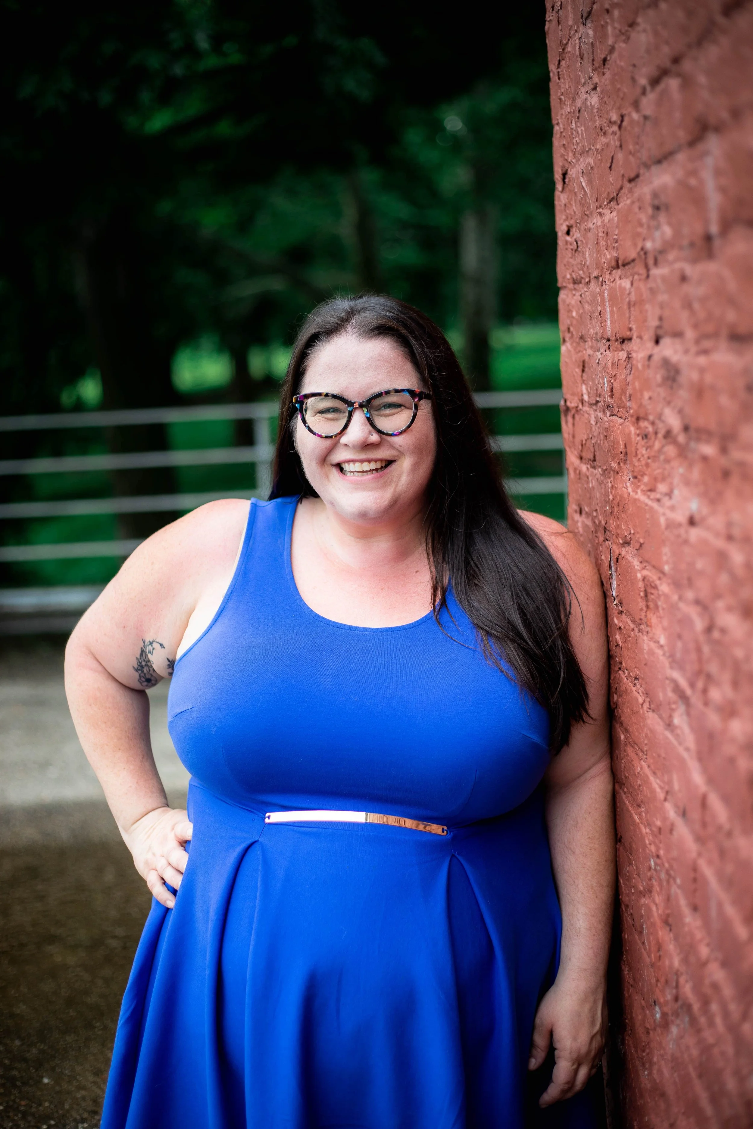 A smiling woman with glasses and long dark hair, wearing a blue sleeveless dress, leaning against a red brick wall outdoors.