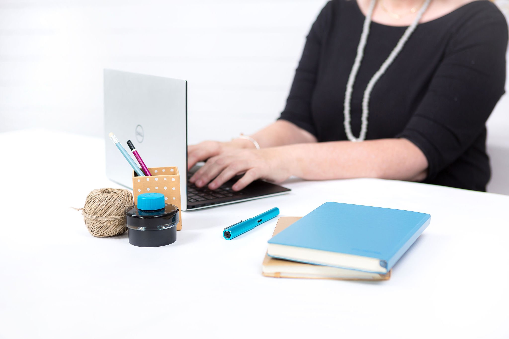 A woman using a laptop at a desk with notebooks, pens, a tape, and a pen holder, against a white background.
