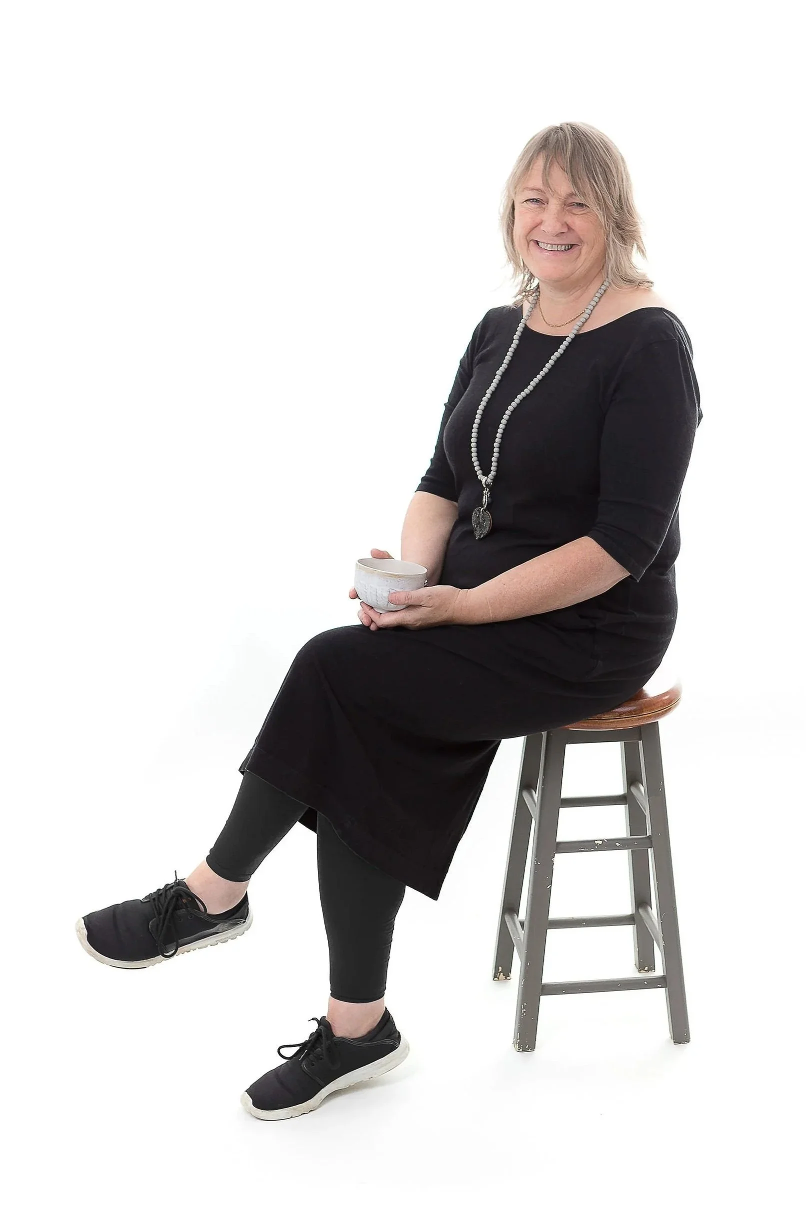 Davina Robertson, smiling and holding a coffee cup and sitting on a stool with a white background