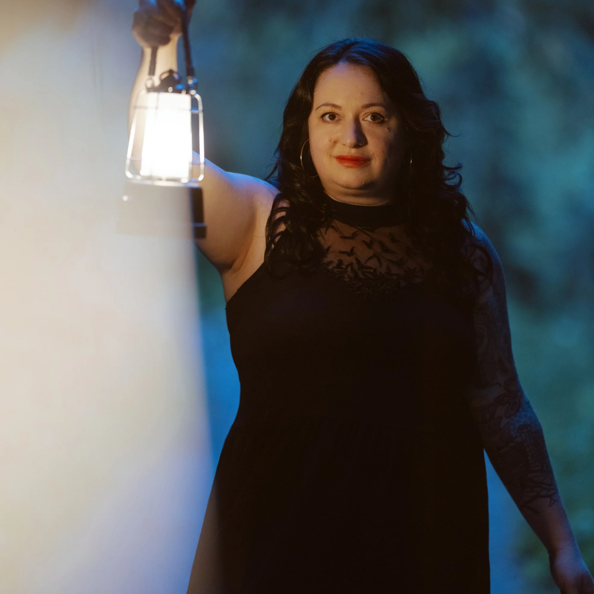 Close-up of a woman with dark, curly hair, wearing red lipstick and hoop earrings, looking at the camera with a neutral expression, in a setting with dark background and a lit lamp on the left side.
