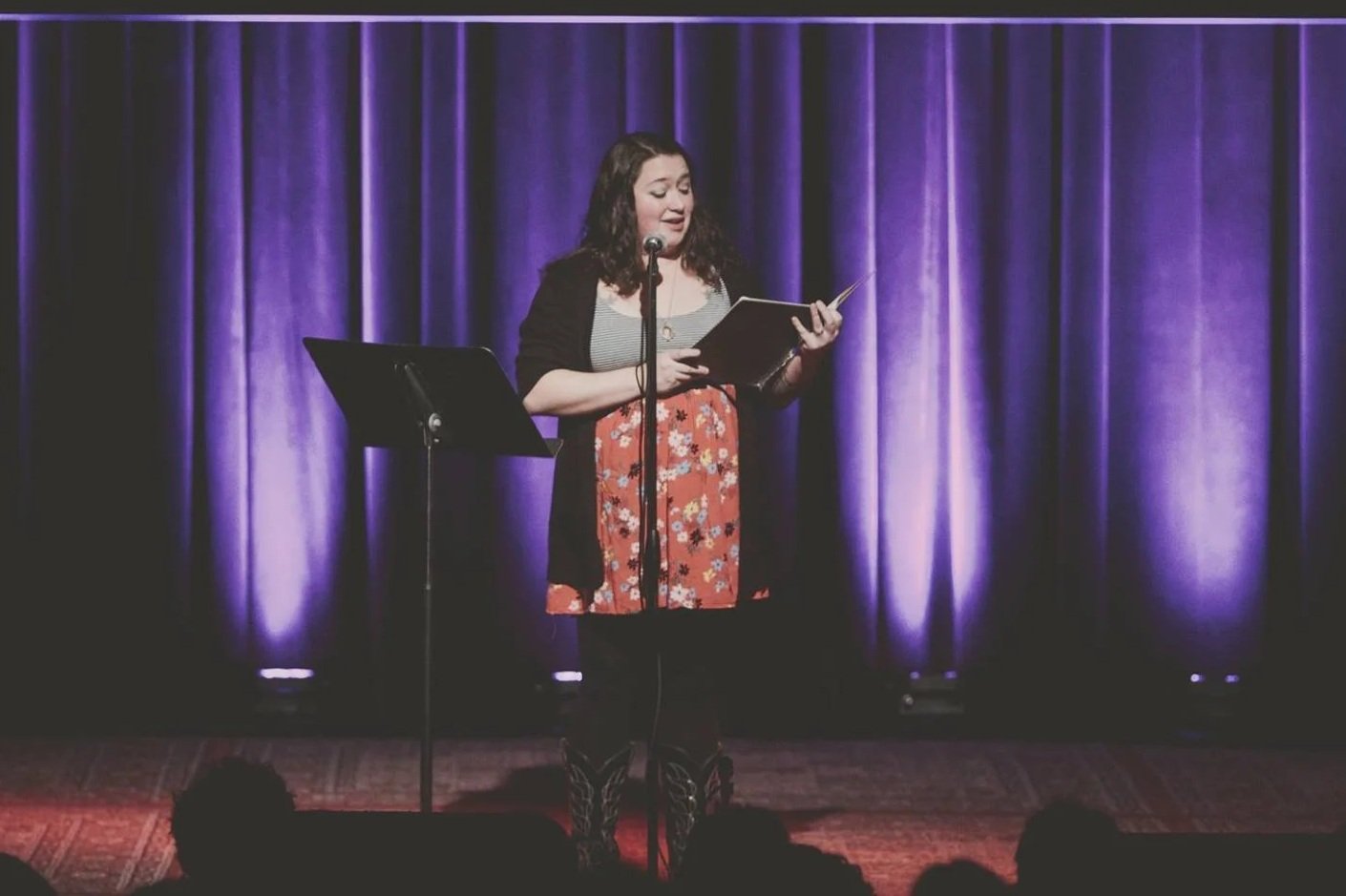 A woman standing on stage, holding a book or script, speaking into a microphone with a purple curtain backdrop.