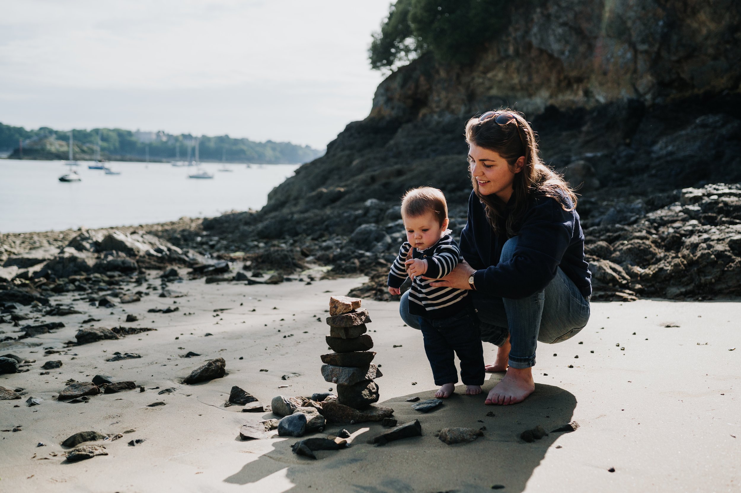 Une femme et un jeune garçon jouent à empiler des pierres sur la plage, avec un arrière-plan de mer, de bateaux et une falaise végétalisée.