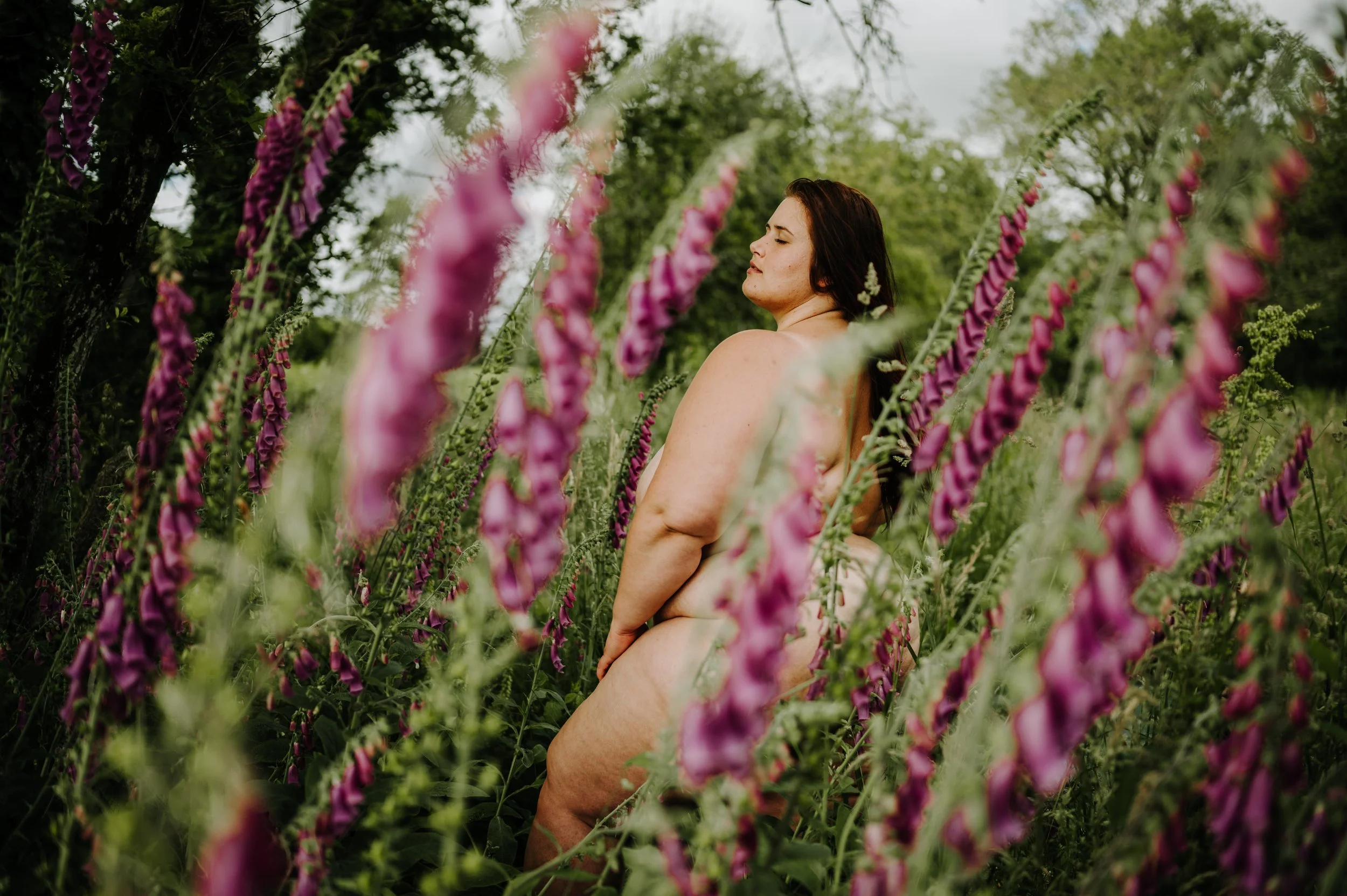 Une femme nue dans un champ de fleurs roses et violettes en plein air, avec des arbres verts en arrière-plan, yeux fermés.